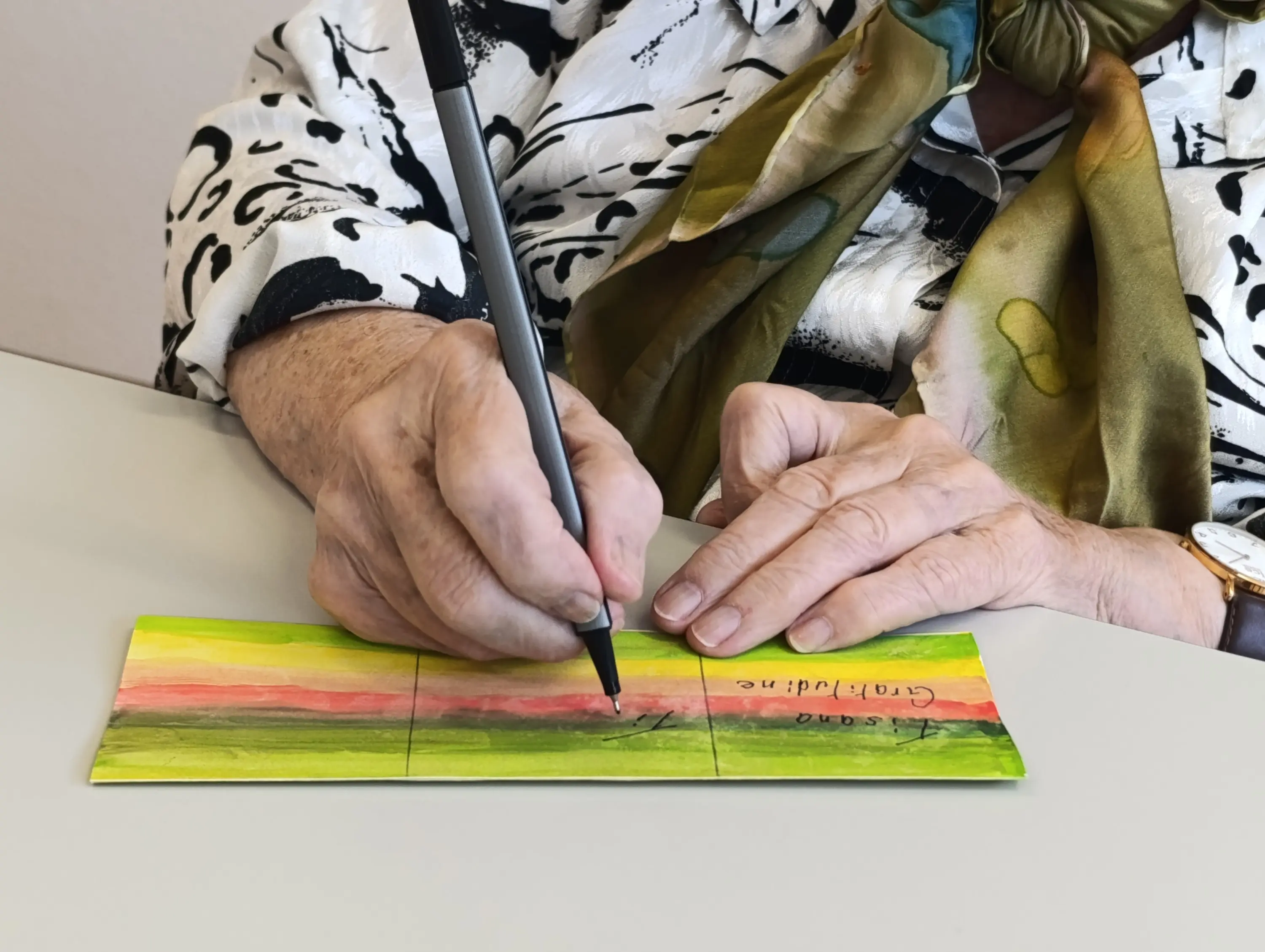 Una persona anziana scrive con un pennarello su un foglio colorato, mentre indossa una camicia a fantasia e una sciarpa in tonalità verde.