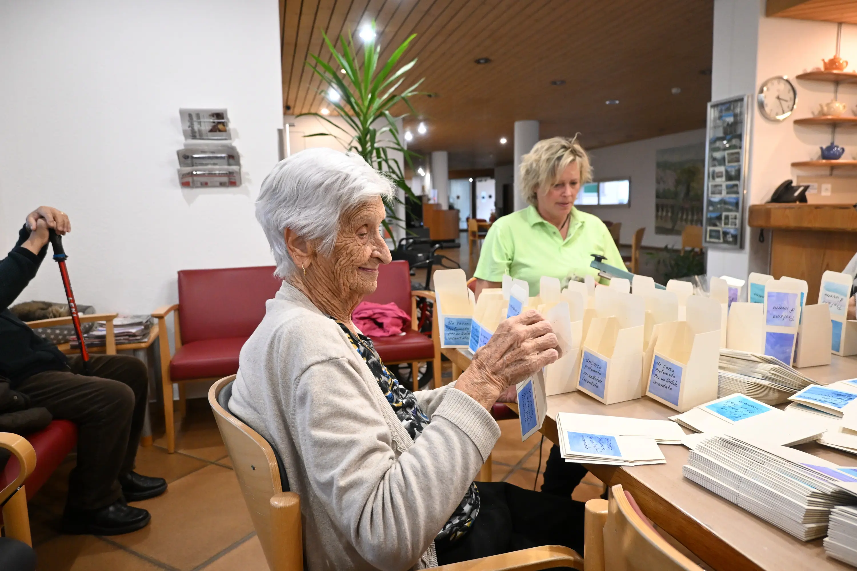 Due donne in un ambiente interno. Una donna anziana sorride mentre tiene in mano un oggetto, mentre l'altra, con i capelli biondi, è seduta accanto a lei, cimentandosi in un'attività. Sul tavolo ci sono diverse scatole e materiale da confe