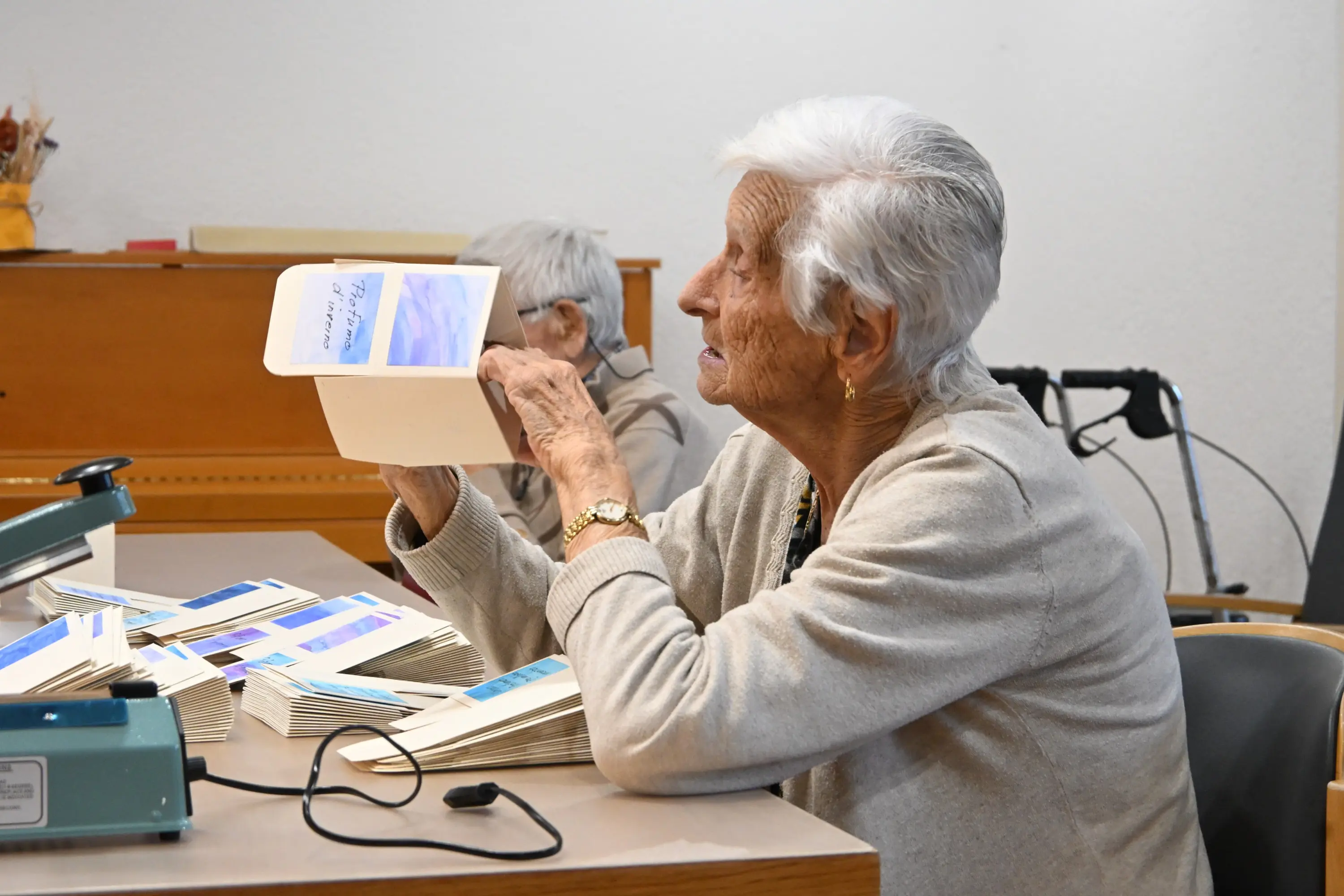 Una donna anziana con capelli bianchi esamina un libro mentre è seduta a un tavolo. Davanti a lei ci sono diversi opuscoli disposti ordinatamente. Un'altra persona anziana è visibile sullo sfondo.