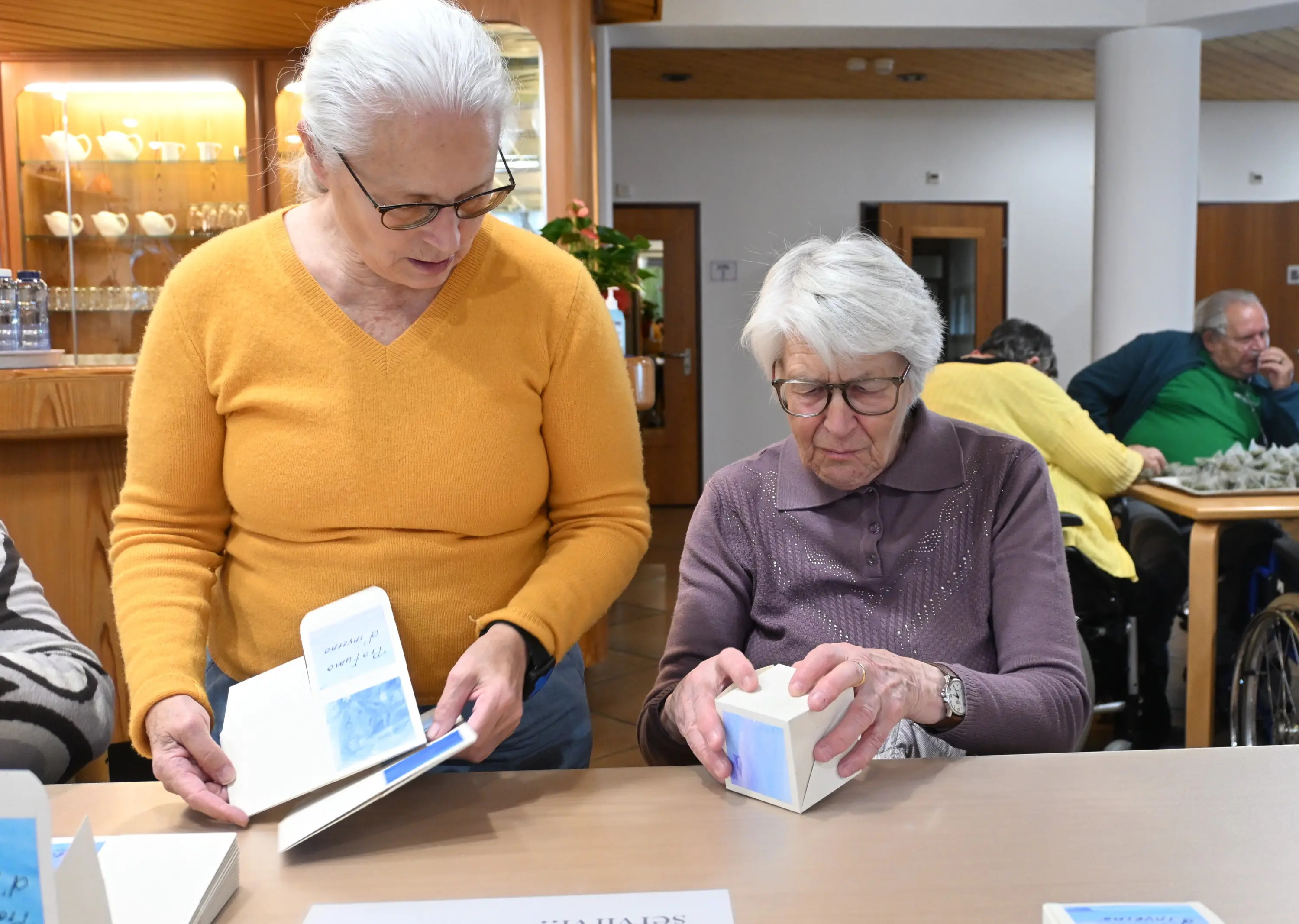 Due donne anziane condividono un momento di attività ricreativa. Una donna in maglione giallo osserva mentre l'altra, con occhiali e un maglione viola, cerca di aprire una scatola. Sul tavolo ci sono vari materiali, inclusi dei
