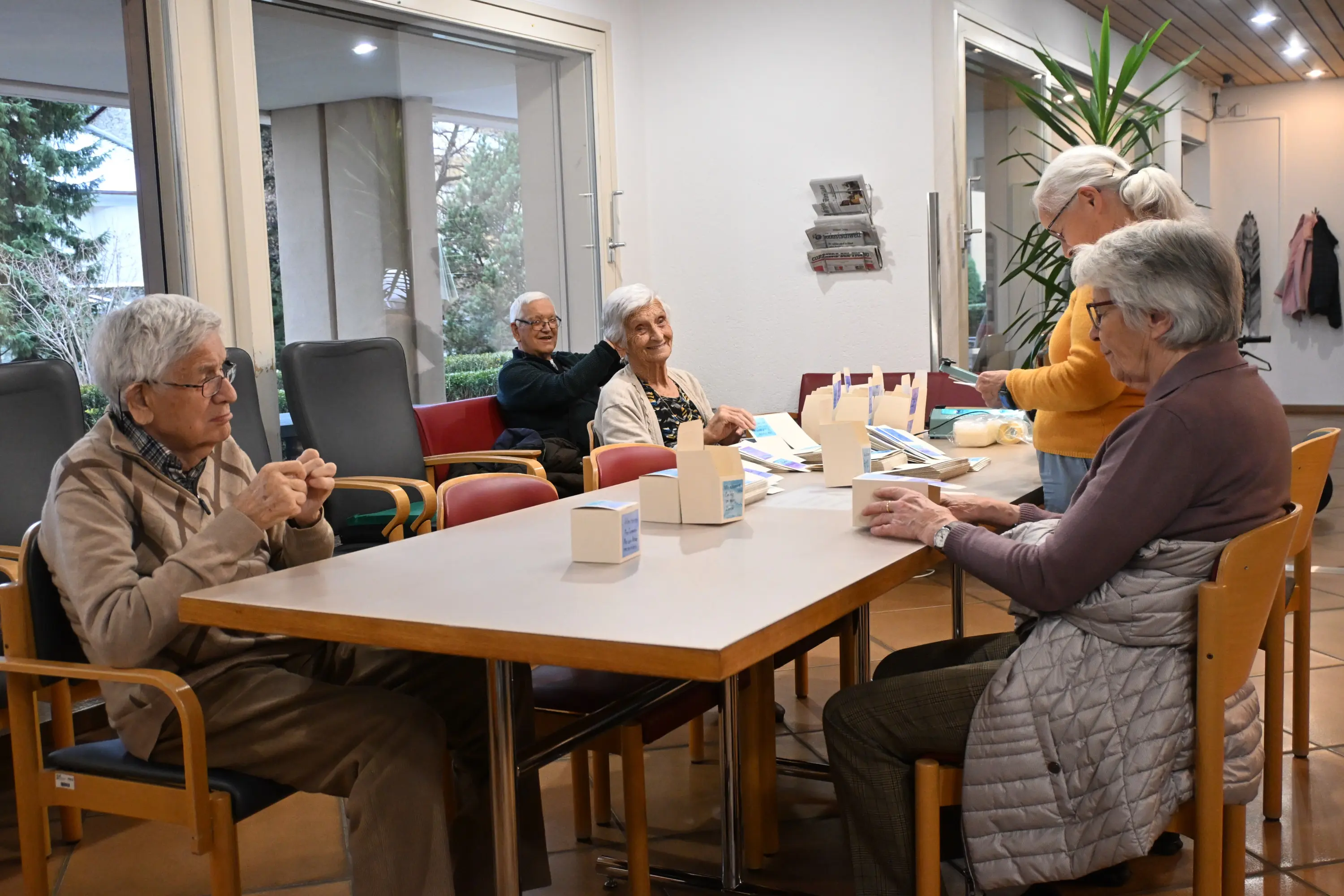 Un gruppo di anziani seduti attorno a un tavolo in una sala comune, mentre alcuni conversano e altri lavorano su attività artigianali. La stanza è luminosa e arredata con piante e sedie colorate.