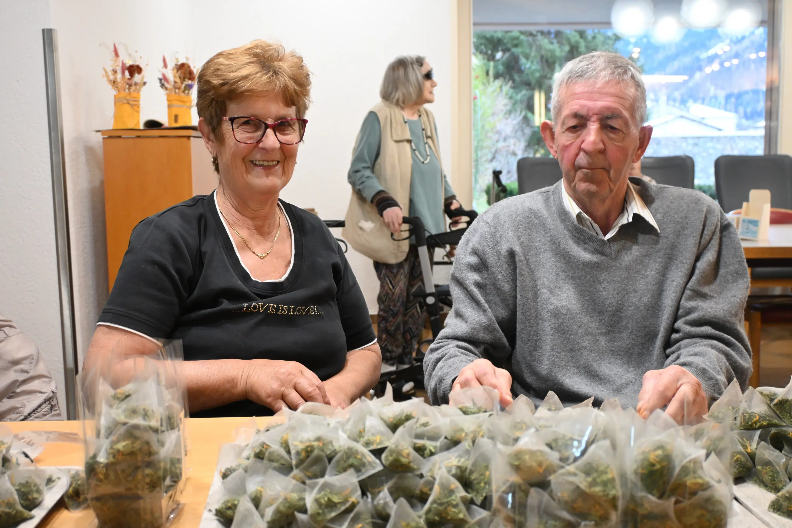 Una donna sorridente e un uomo serio siedono a un tavolo, circondati da sacchetti di erbe. Sullo sfondo, una donna anziana cammina con un bastone.