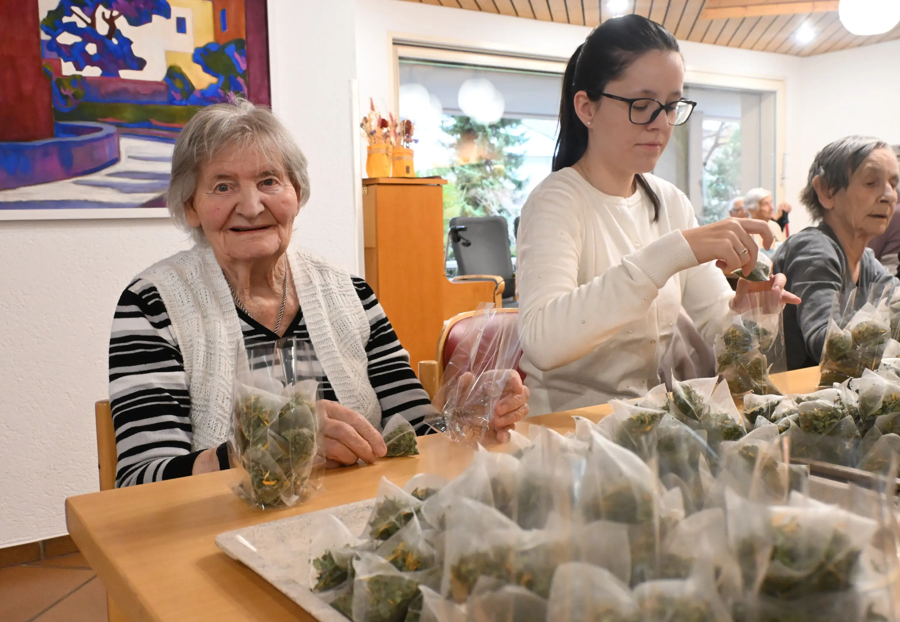 Un'anziana sorridente e una giovane donna lavorano insieme in un centro, confezionando sacchetti di erbe su un tavolo. Sullo sfondo si intravedono altre anziane. L'atmosfera è conviviale e laboriosa.