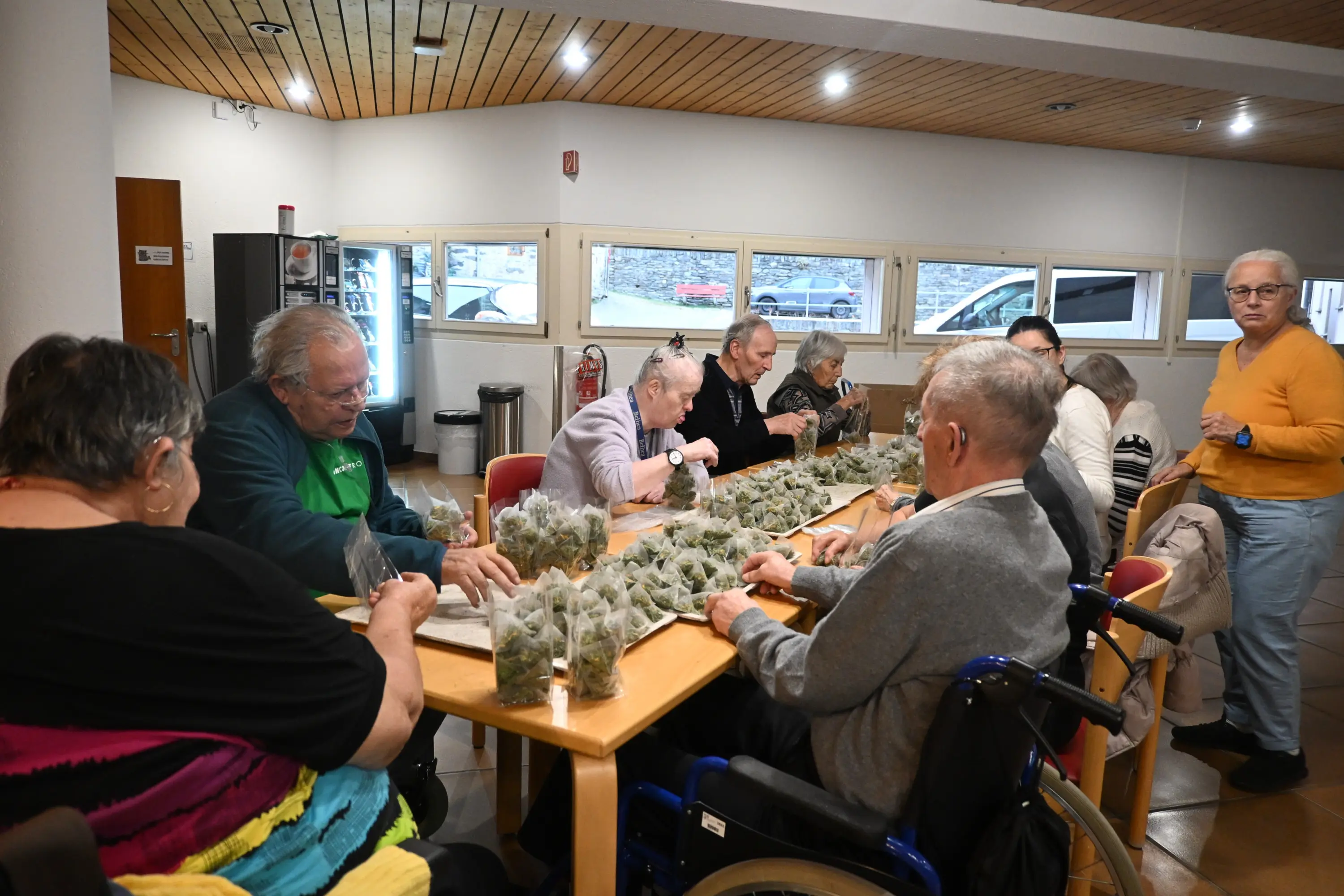 Un gruppo di persone anziane, alcune su sedie a rotelle, è seduto attorno a un tavolo mentre lavorano insieme a un'attività manuale. Sul tavolo ci sono numerosi sacchetti di plastica con contenuti verdi, probabilmente erbe o piant