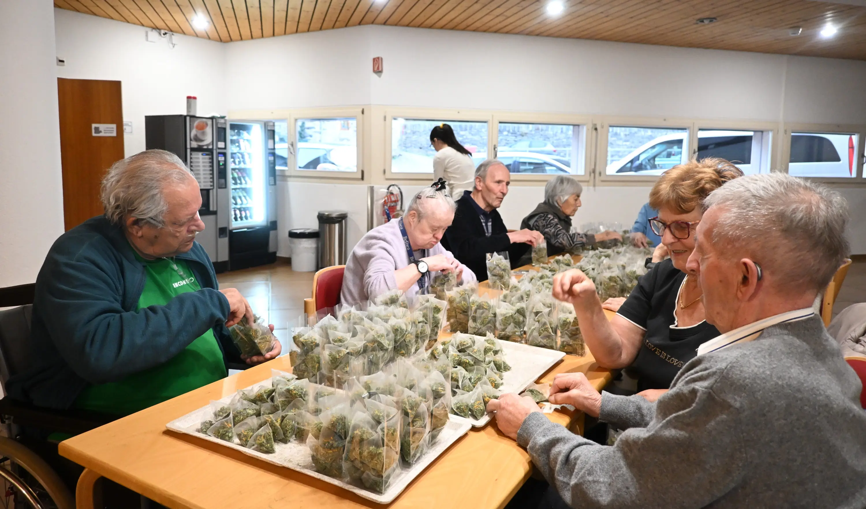 Un gruppo di persone anziane, sedute a un tavolo, sta confezionando sacchetti di erbe. Alcuni di loro sorridono e conversano, mentre altri si concentrano sul loro compito. L'ambiente è luminoso, con finestre che mostrano un