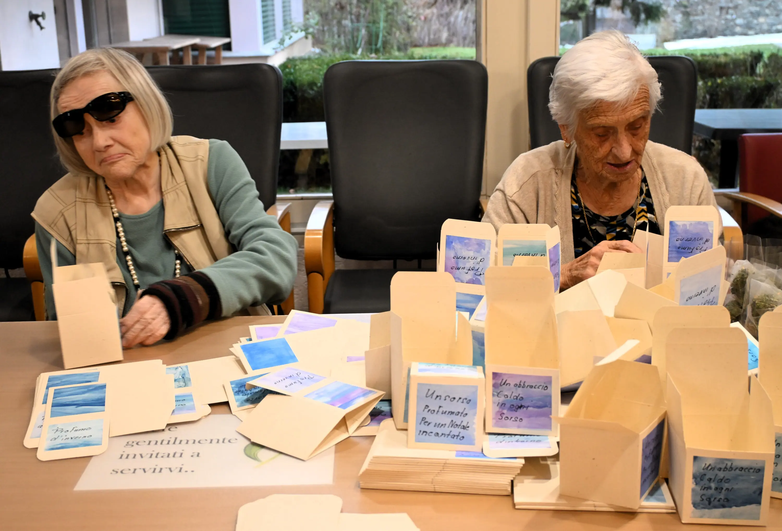 Due donne anziane sedute a un tavolo, circondate da scatoline vuote e cartoncini colorati. Una donna indossa occhiali scuri e sorride mentre l'altra scrive su un cartoncino. Sul tavolo ci sono delle scritte visibili.