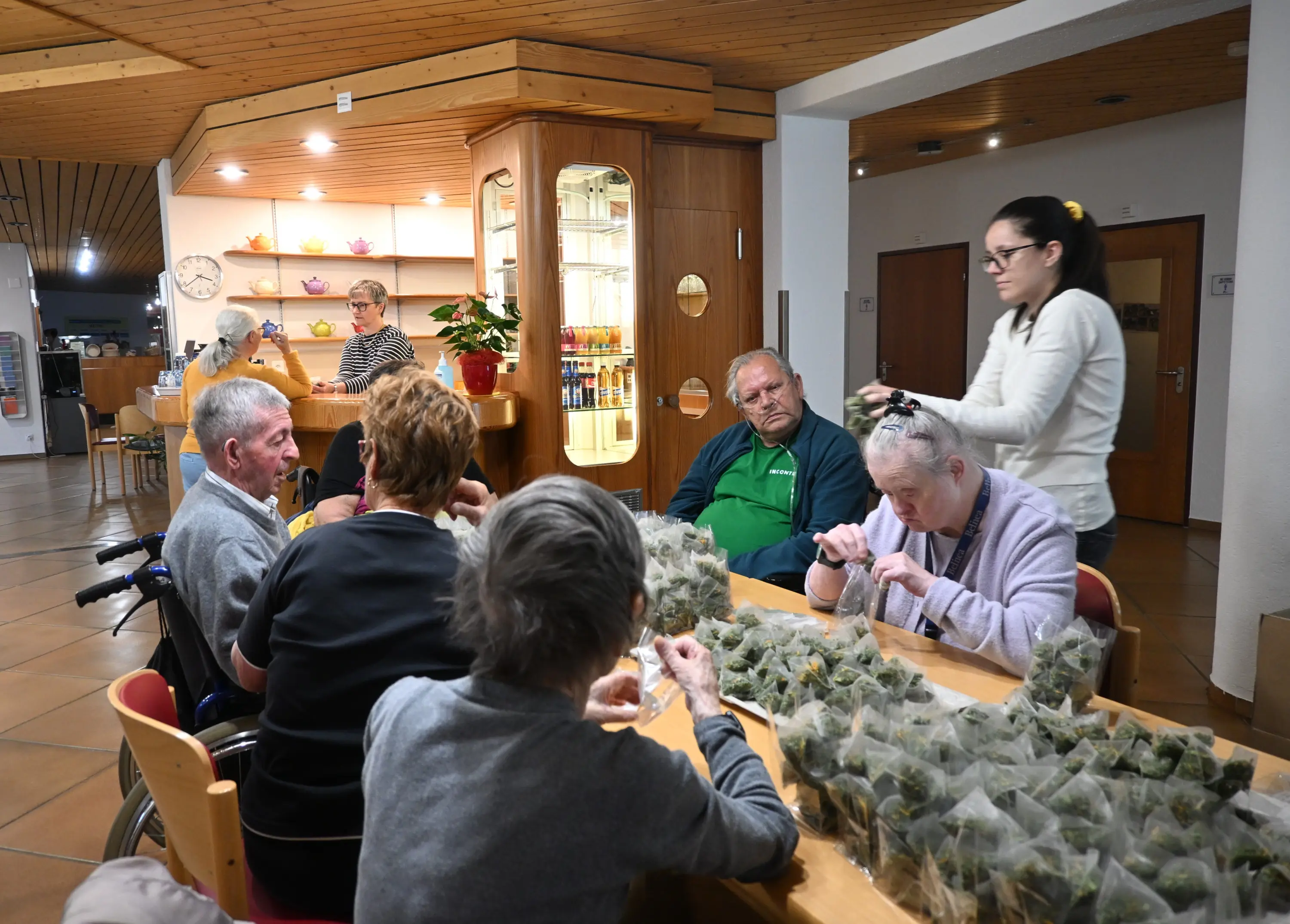 Un gruppo di persone anziane si riunisce attorno a un tavolo, mentre un'operatrice distribuisce delle borse di plastica contenenti probabilmente erbe o dolci. Alcuni anziani chiacchierano, mentre altri sono concentrati sulle loro attività. Sul
