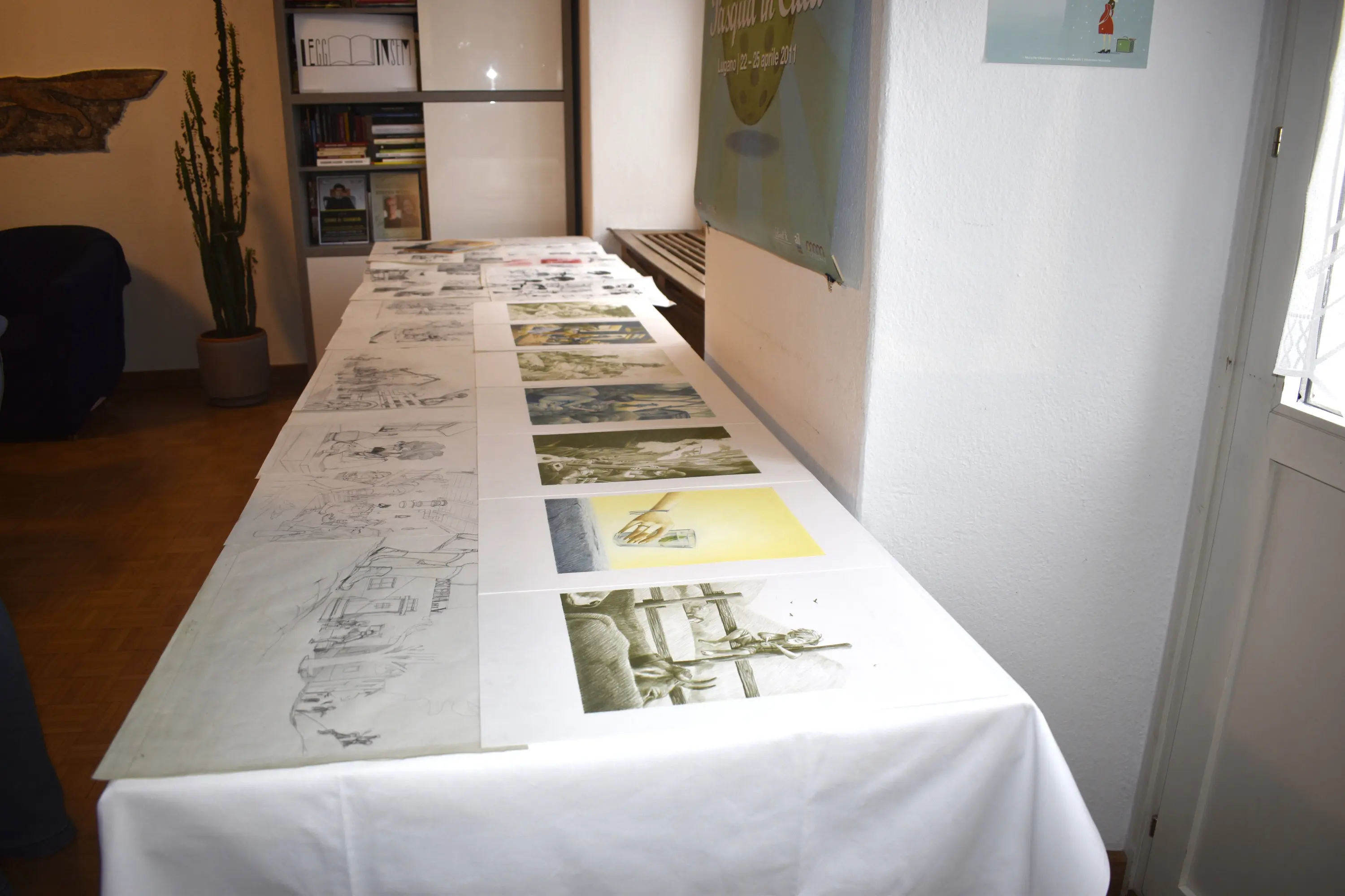 Un tavolo coperto da una tovaglia bianca, adagiato con disegni e illustrazioni di varie dimensioni, in un ambiente interno con una pianta e una finestra sullo sfondo.