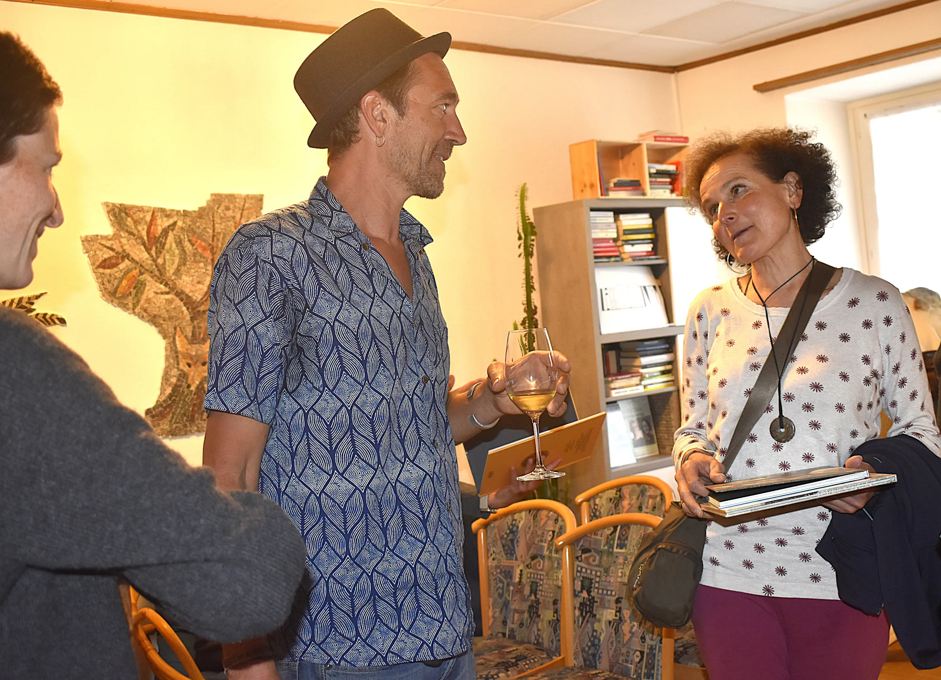 Due persone sono in conversazione in una stanza, mentre un'altra persona osserva. Uno degli interlocutori indossa un cappello e tiene un bicchiere in mano. La decorazione della stanza include libri sugli scaffali e un'opera d'arte sulla parete.