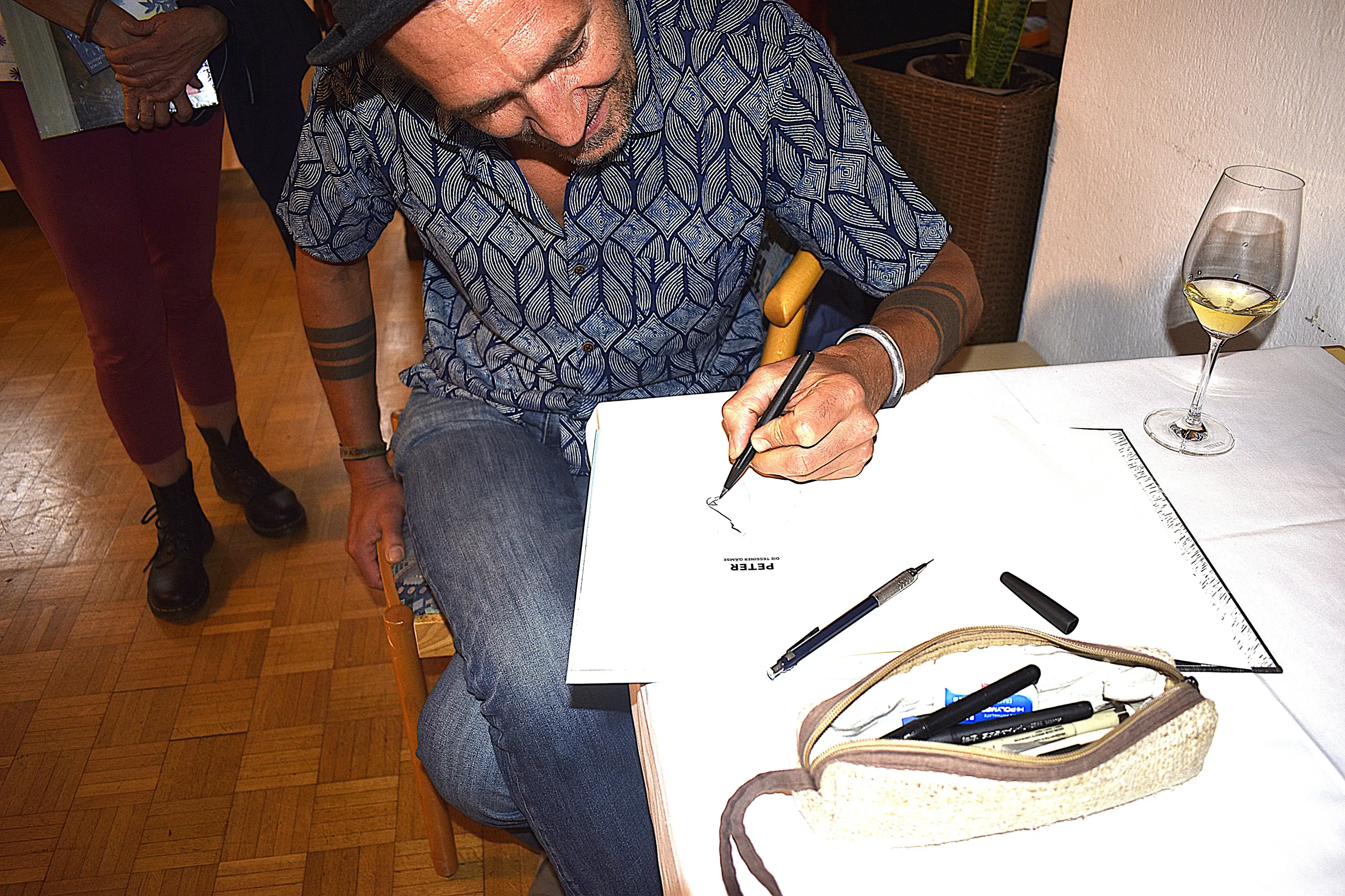 Uomo seduto che disegna su un quaderno con una penna, sorridendo. Ha un cappello e una camicia a motivi geometrici. Sul tavolo sono visibili un bicchiere con una bevanda e una borsa con penne e matite.