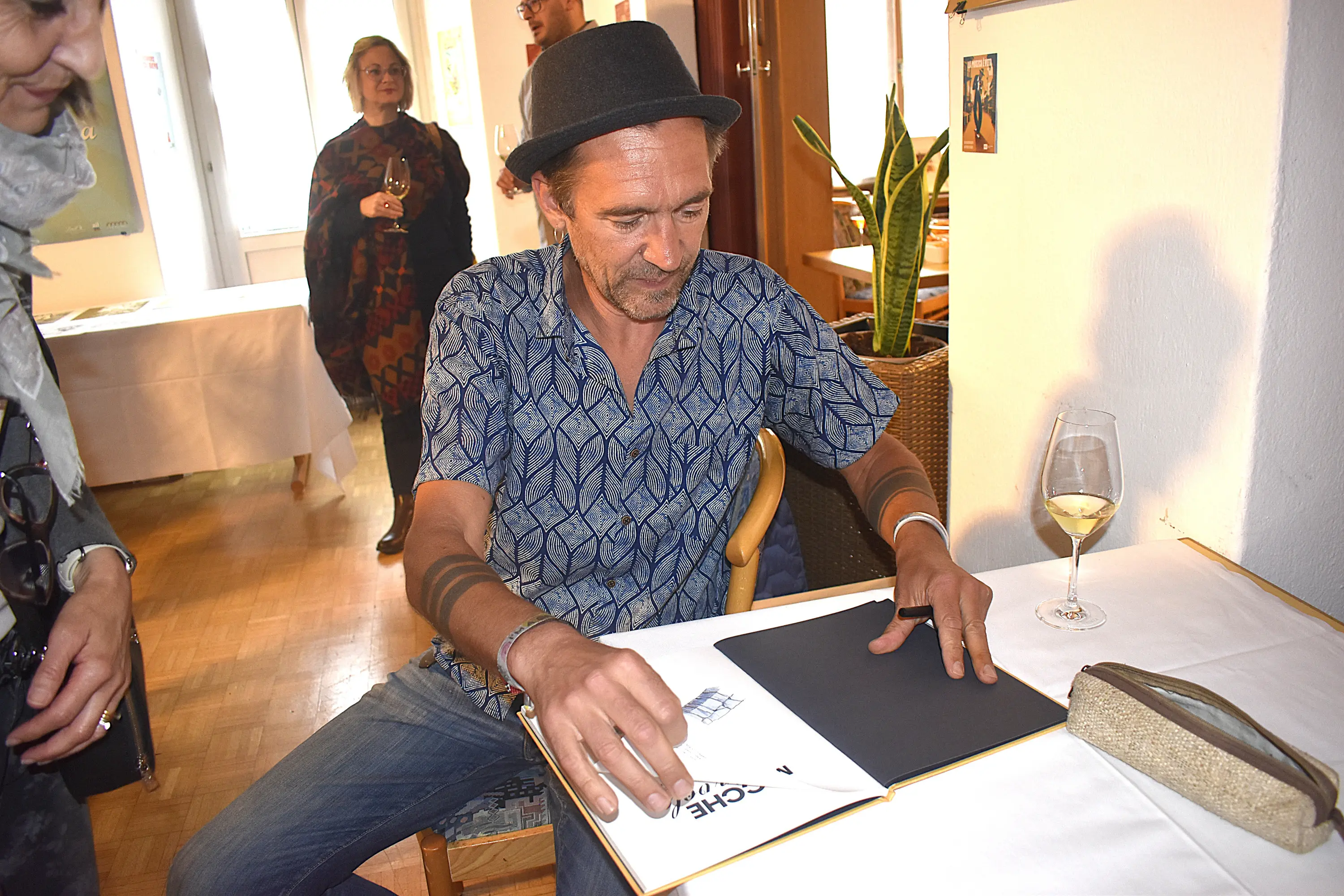 Uomo con cappello che sfoglia un libro in un ambiente informale, circondato da persone. Sul tavolo ci sono un bicchiere di vino bianco e un oggetto decorativo.