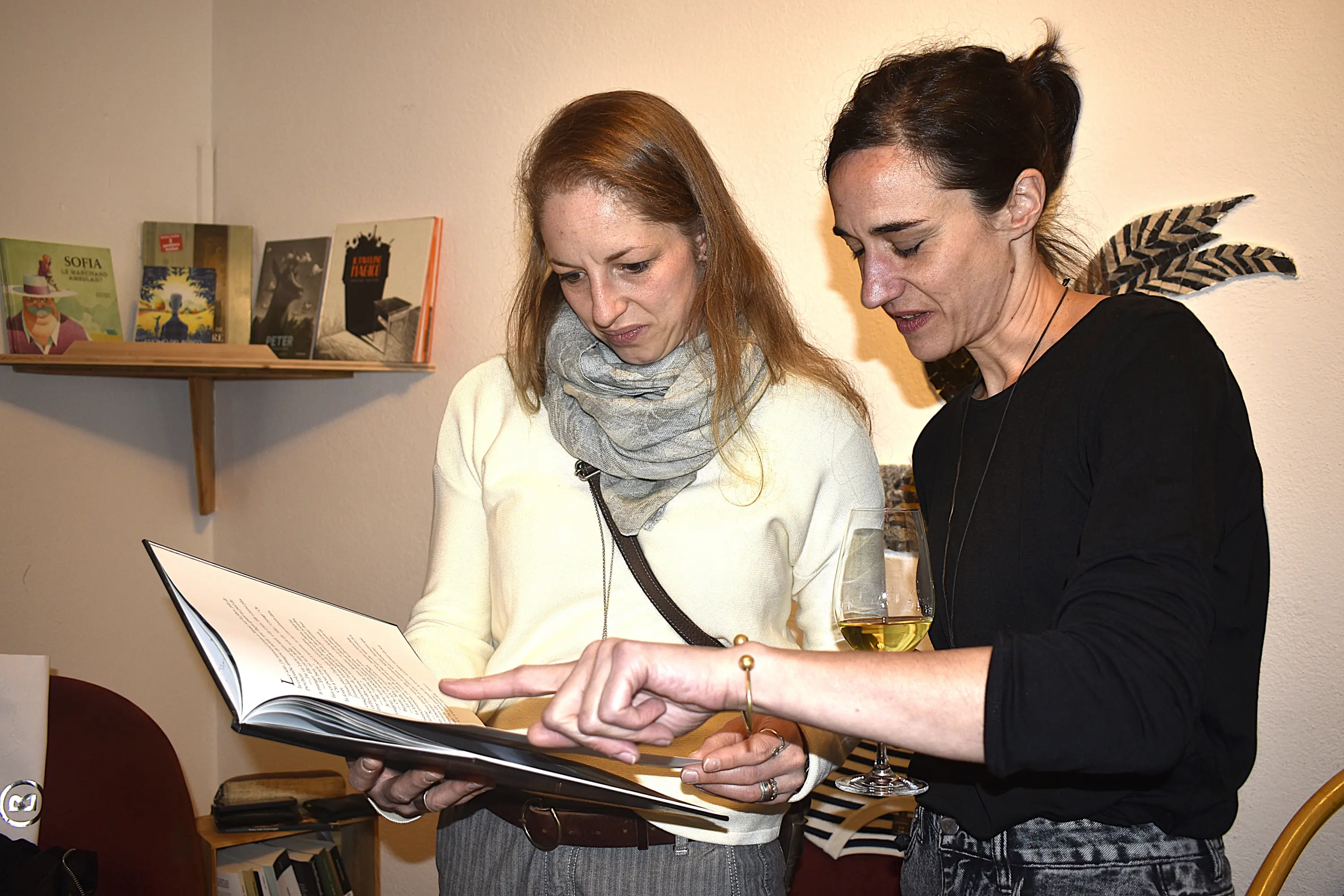 Due donne sfogliano un grande libro mentre discutono tra loro. Una donna indossa una sciarpa grigia e ha i capelli lunghi e castani, mentre l'altra ha i capelli scuri raccolti e indossa una maglietta nera. Sullo sfondo, si intravedono libri su una mensola.