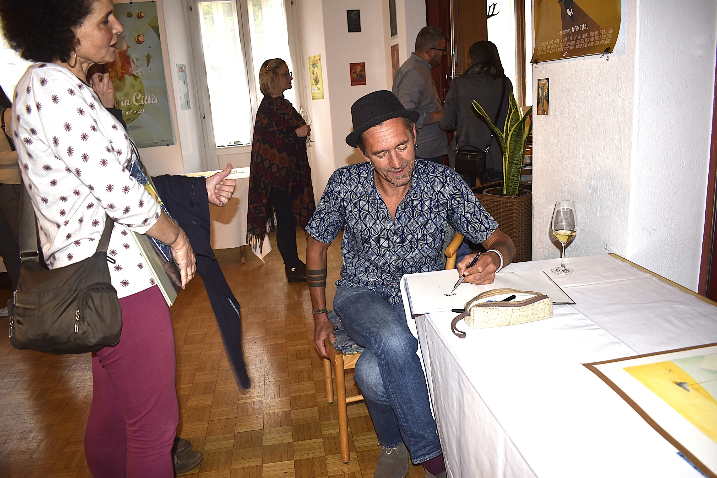 Un uomo elegante, con un cappello nero, scrive su un quaderno mentre siede a un tavolo. Una donna, in secondo piano, osserva e tiene un libro in mano. L'ambiente è decorato con opere d'arte e ci sono altre persone che interagiscono nel locale. Una coppa di vino bianco è appoggiata sul tavolo.