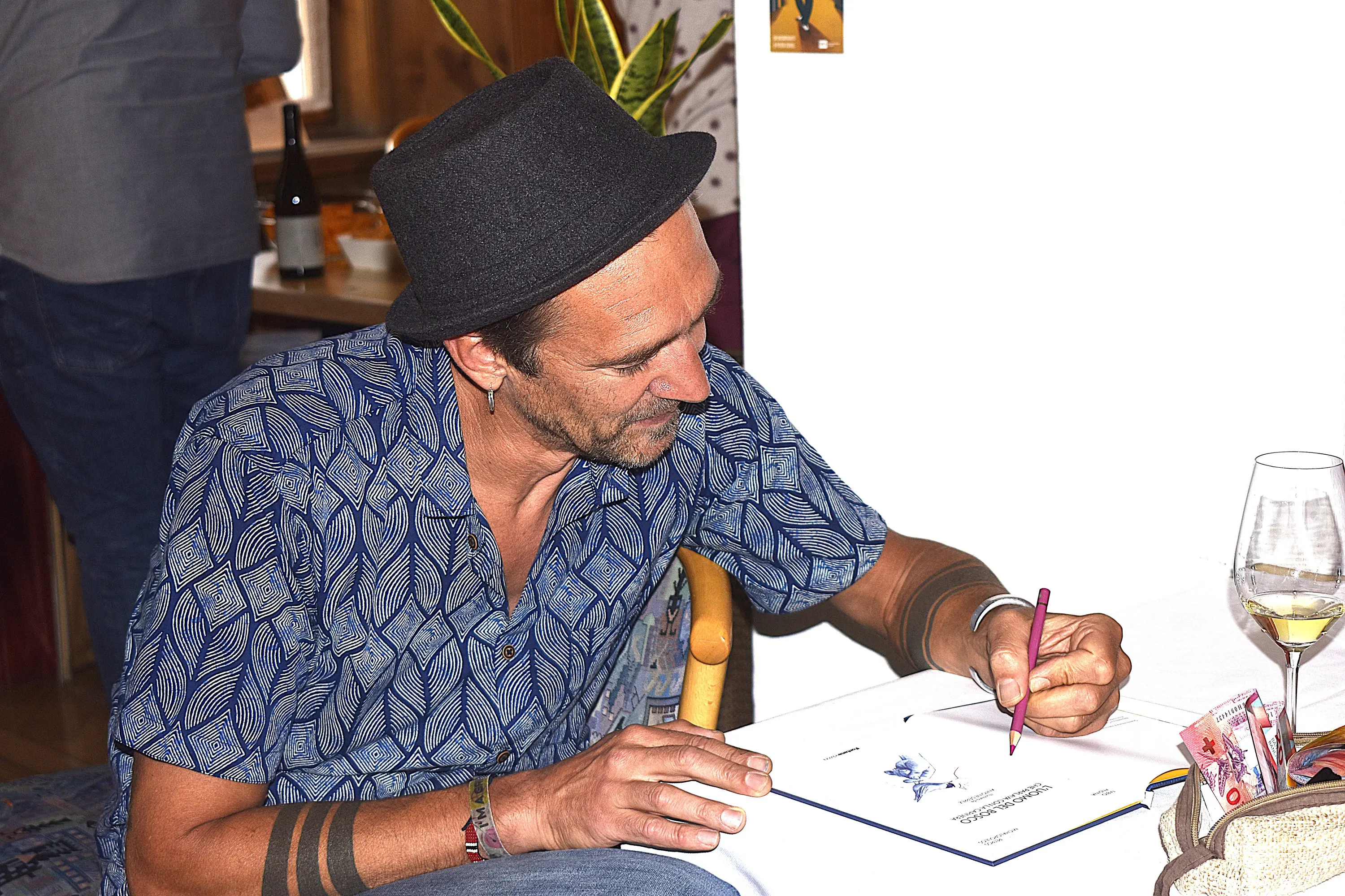 Uomo con cappello a cilindro disegna su un foglio mentre è seduto a un tavolo. Indossa una camicia a maniche corte con un motivo blu e tiene in mano una matita. Sul tavolo c'è un bicchiere di vino e altri oggetti.