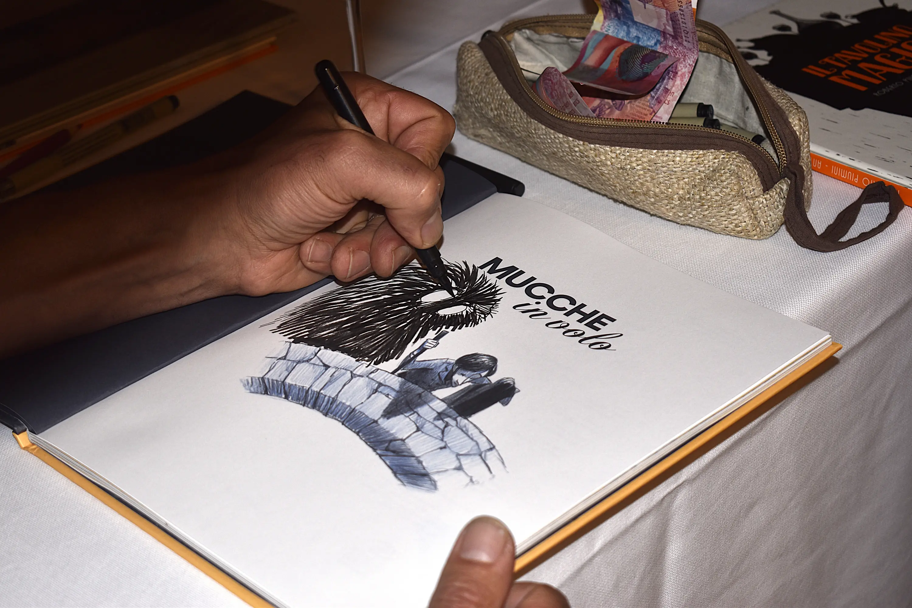 Una mano che disegna su una pagina di un libro aperto. Il disegno rappresenta una figura stilizzata con spine e il titolo "Mucche in volo" scritto sopra. Accanto, c'è una borsa di stoffa e un altro libro.