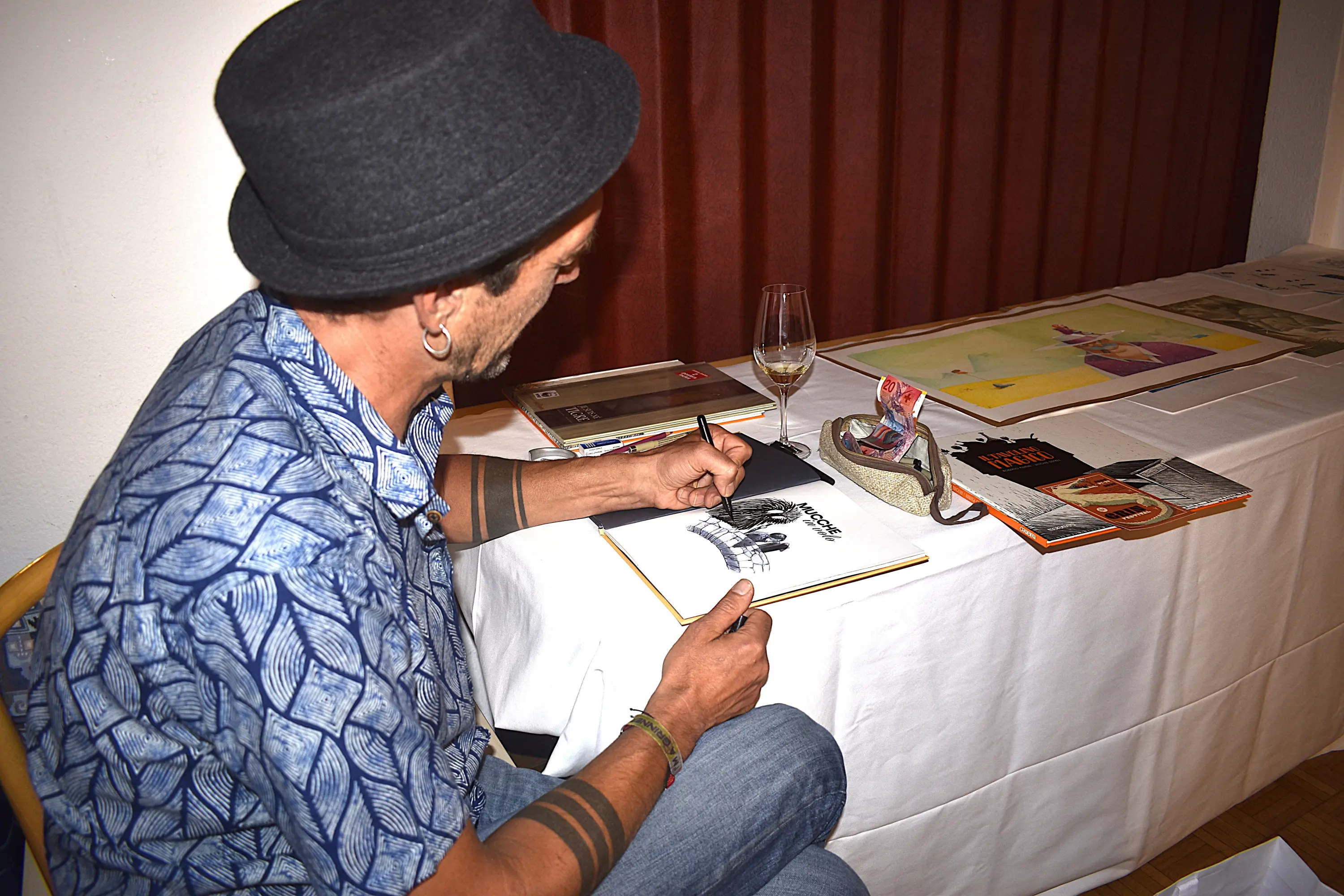 Un uomo con un cappello di feltro disegna su un blocco di carta mentre tiene un bicchiere di vino. Sul tavolo si trovano vari materiali artistici e una borsa.