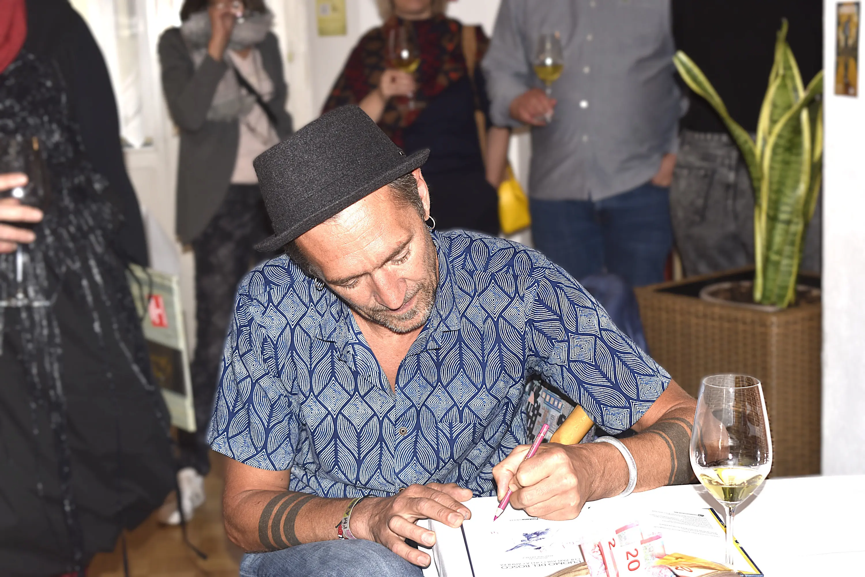 Un uomo in camicia con un motivo geometrico e cappello di feltro scrive su un foglio mentre tiene un bicchiere di vino. Sullo sfondo si intravedono altre persone in un ambiente festoso.