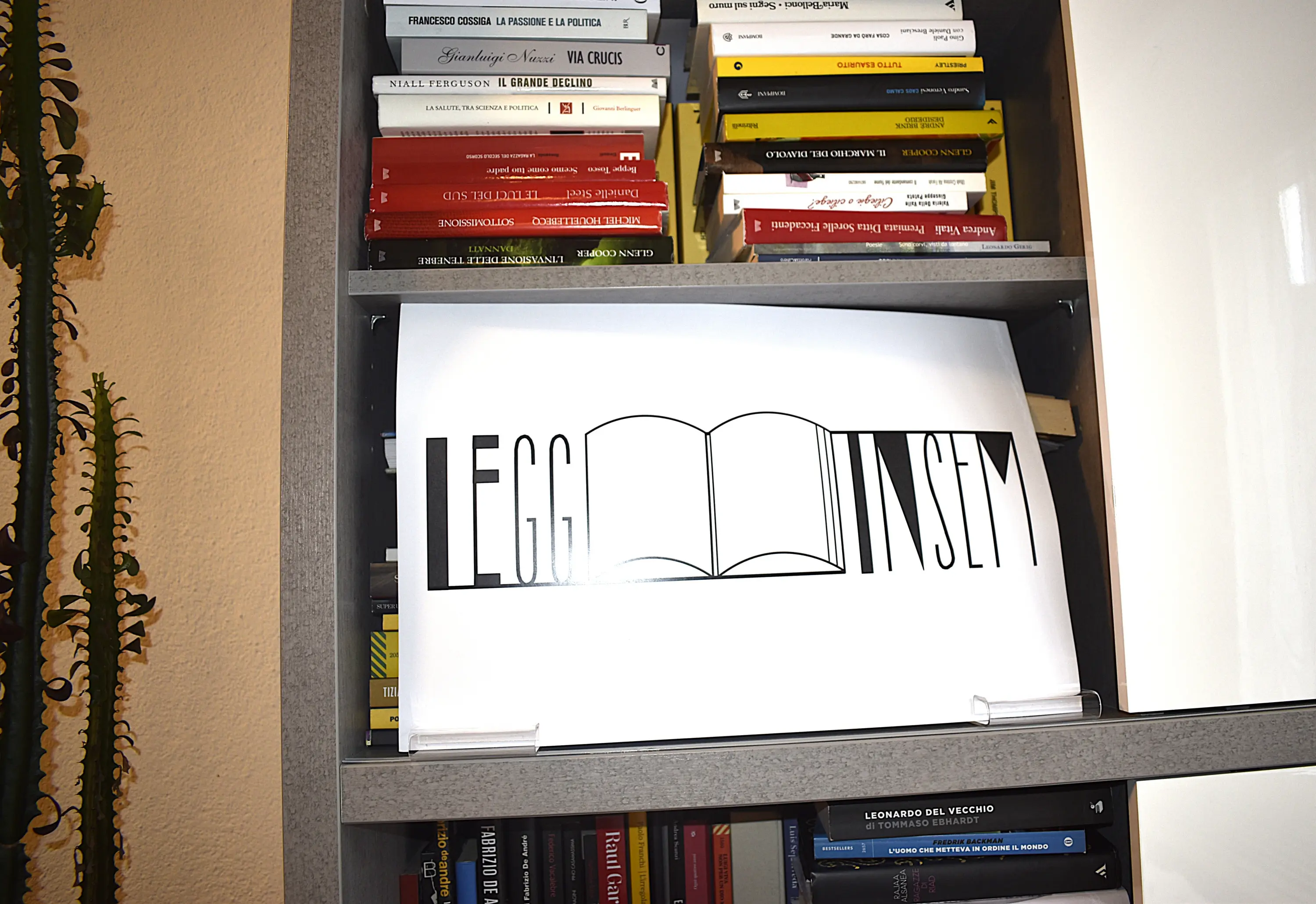 Un cartello con la scritta "LEGGI INSIEME" su sfondo bianco, posizionato su una mensola di libri. Sullo sfondo, si vedono libri disposti ordinatamente.