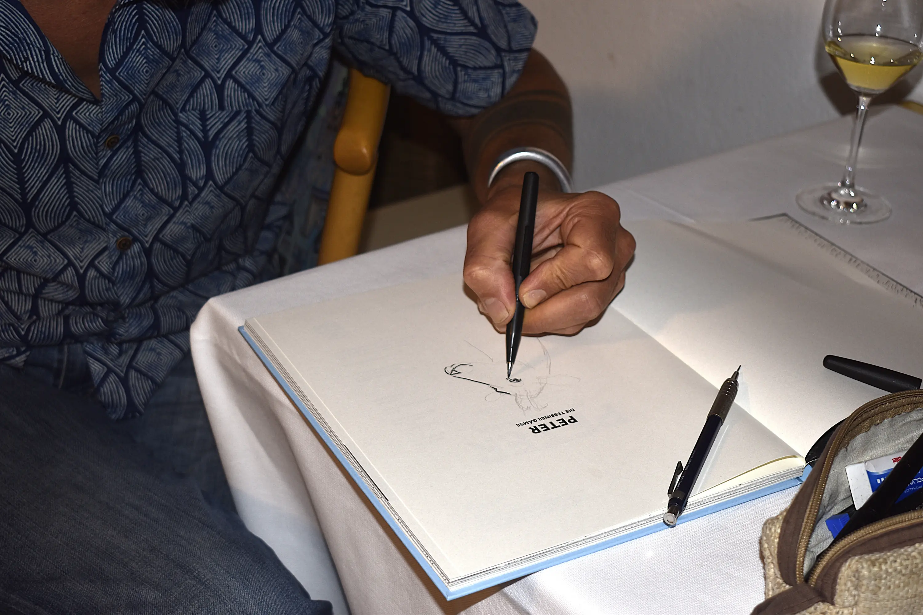 Una mano disegna su un foglio aperto con una penna, mentre un bicchiere di vino è appoggiato su un tavolo. Sul foglio si legge il nome "PETER".