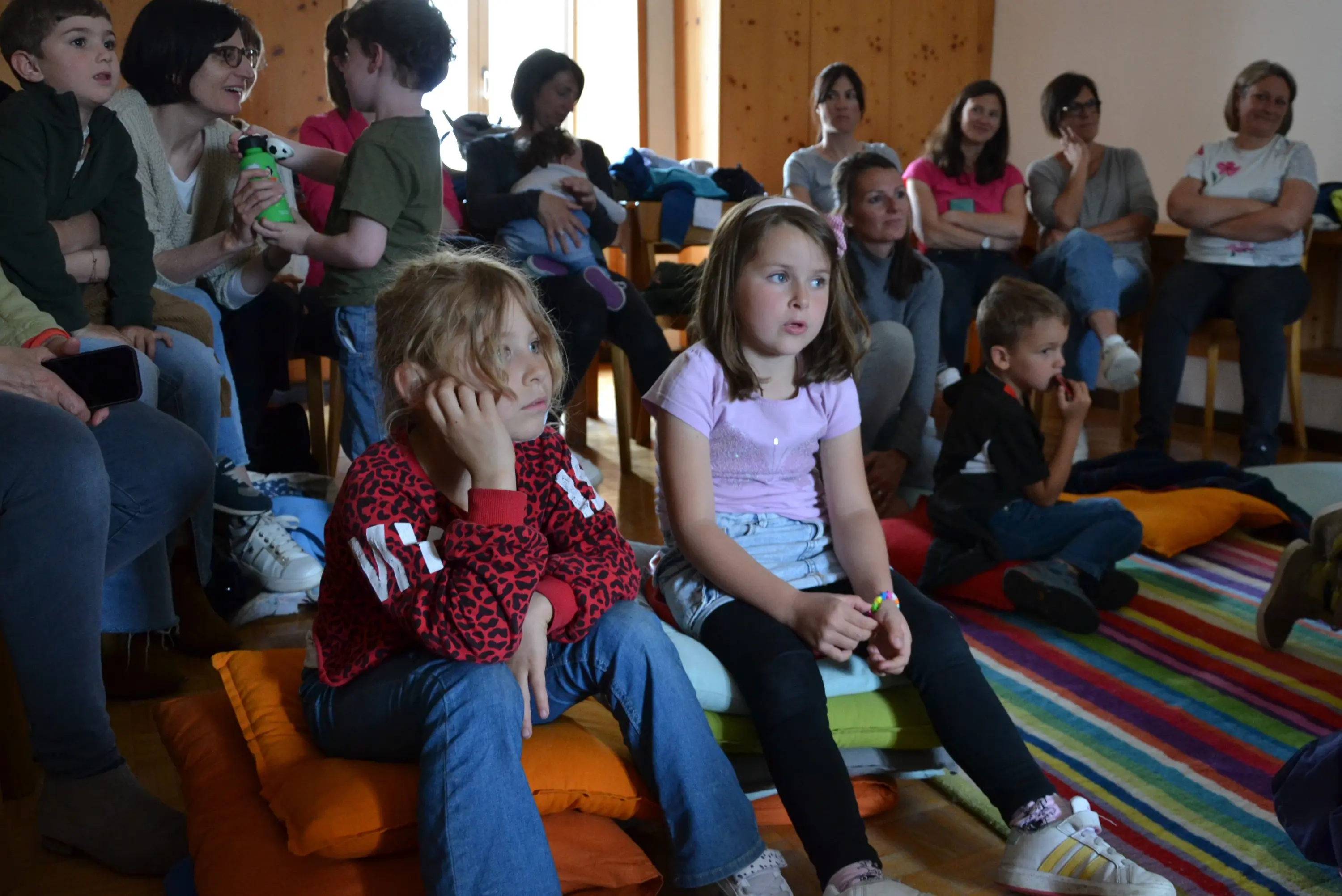 Un gruppo di bambini e adulti seduti su cuscini colorati in una stanza. Due bambine in primo piano osservano attentamente, mentre altri partecipanti sono visibili sullo sfondo, alcuni sorridendo.