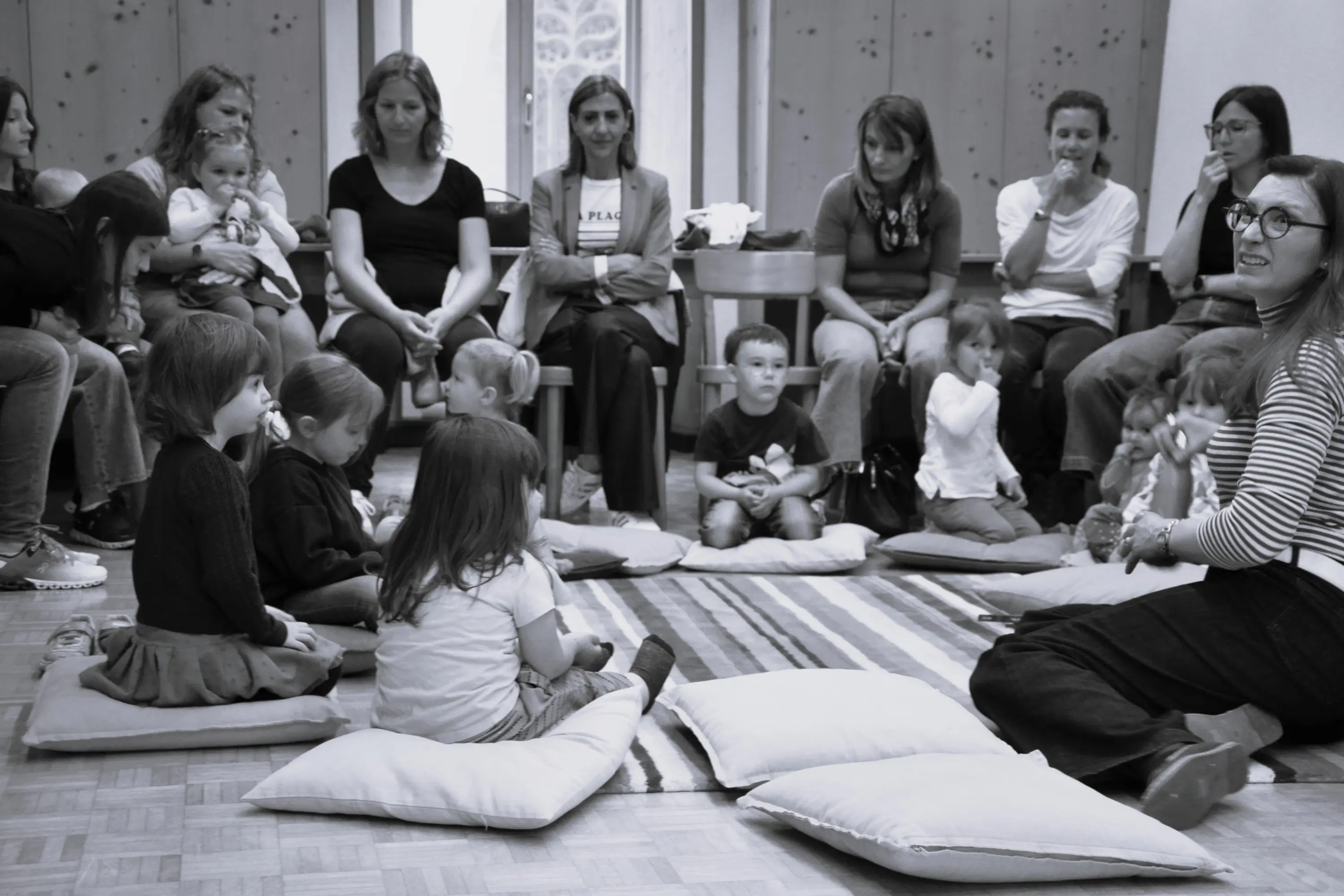 Gruppo di adulti e bambini seduti su cuscini in una stanza. Alcuni adulti tengono in braccio i bambini mentre altri li osservano. Una donna, seduta davanti al gruppo, sta raccontando una storia ai bambini.