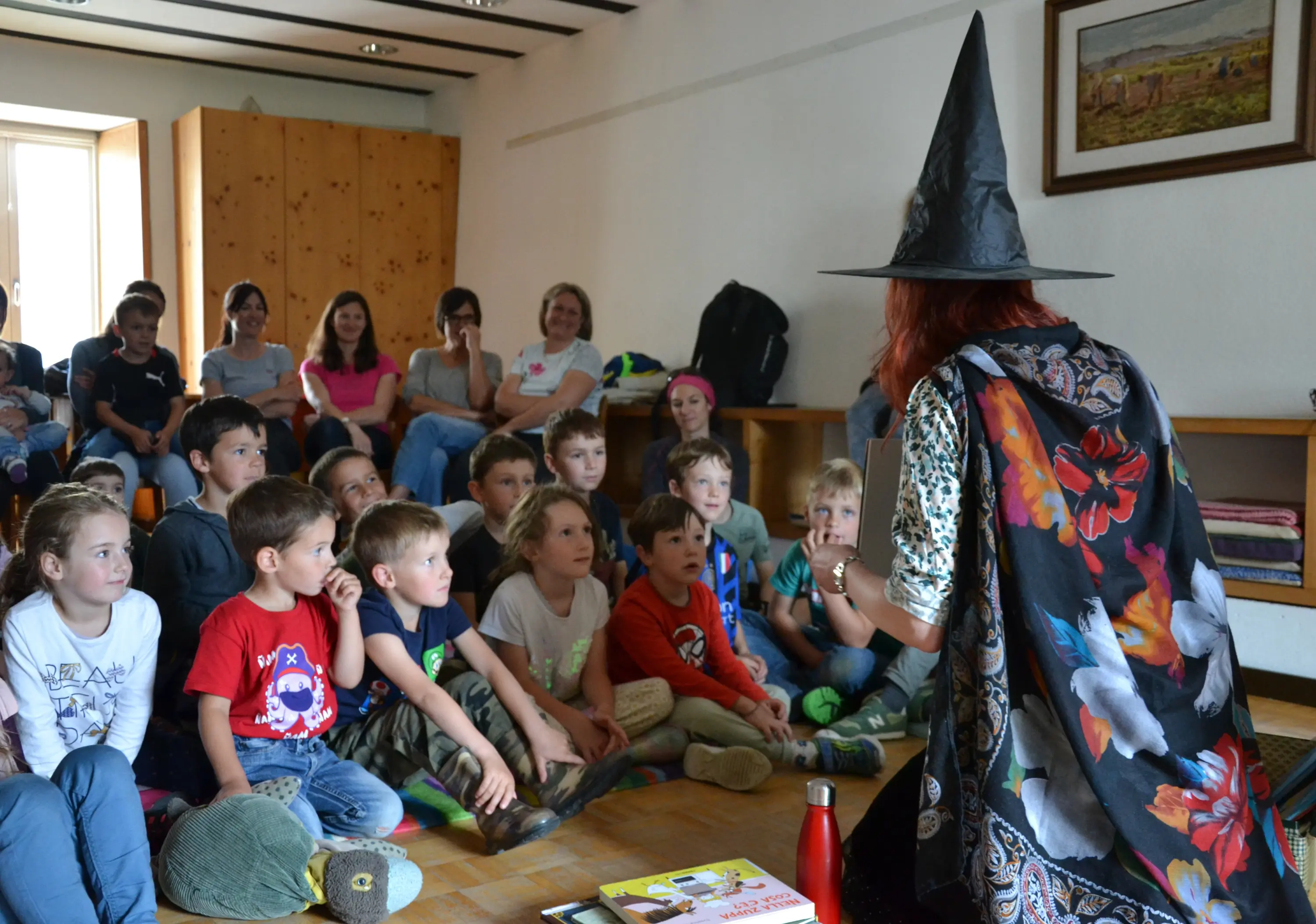 Un gruppo di bambini ascolta attentamente una narratrice travestita da strega in una stanza luminosa. Alcuni adulti sorridono sullo sfondo, mentre i bambini sono seduti in cerchio, incuriositi.