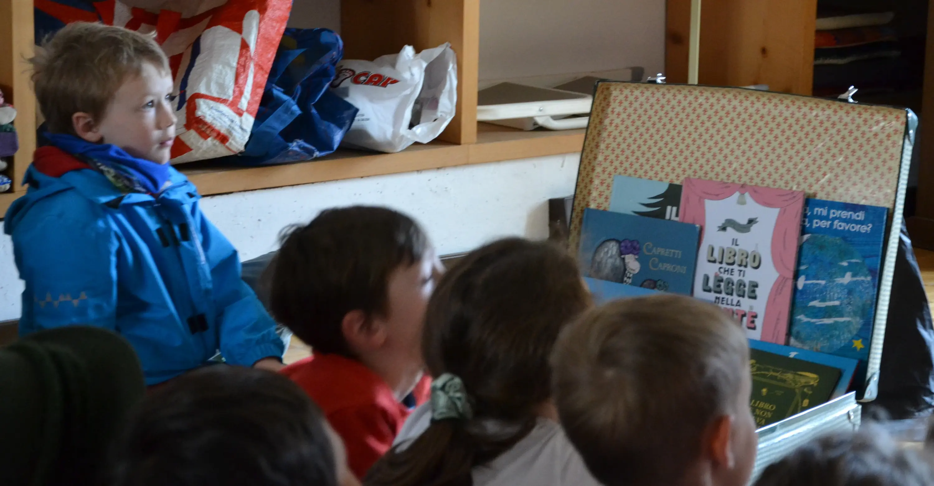 Un bambino con giacca blu osserva attentamente i libri in una valigia aperta. Dietro di lui, un gruppo di bambini è seduto, concentrato sulla presentazione.