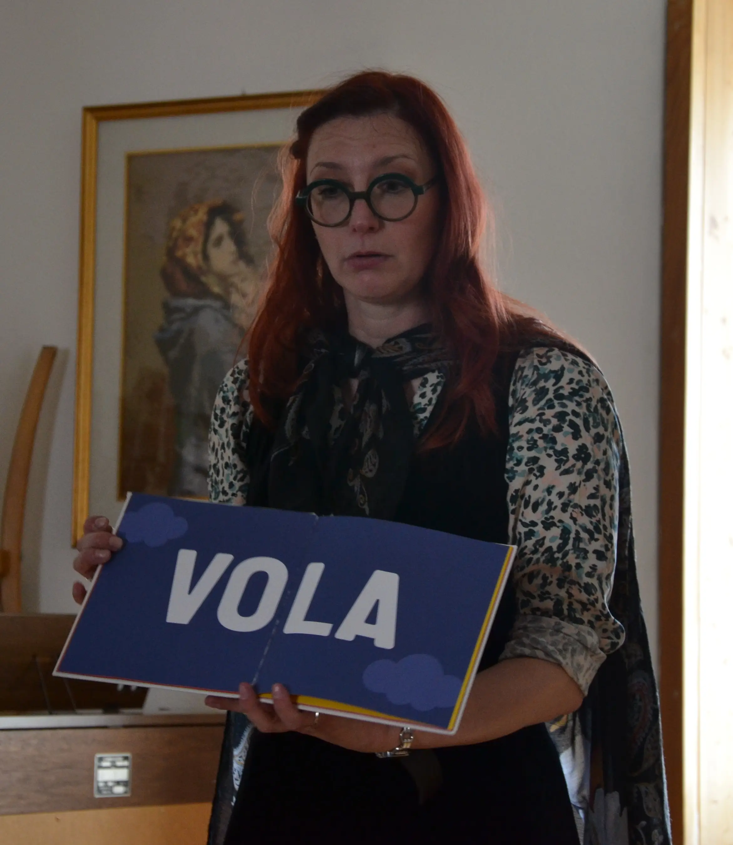 Donna con occhiali tondi e capelli rossi, tiene in mano un cartello blu con la scritta "VOLA". Indossa una camicia a fiori e un foulard. Sullo sfondo si nota un'opera d'arte.