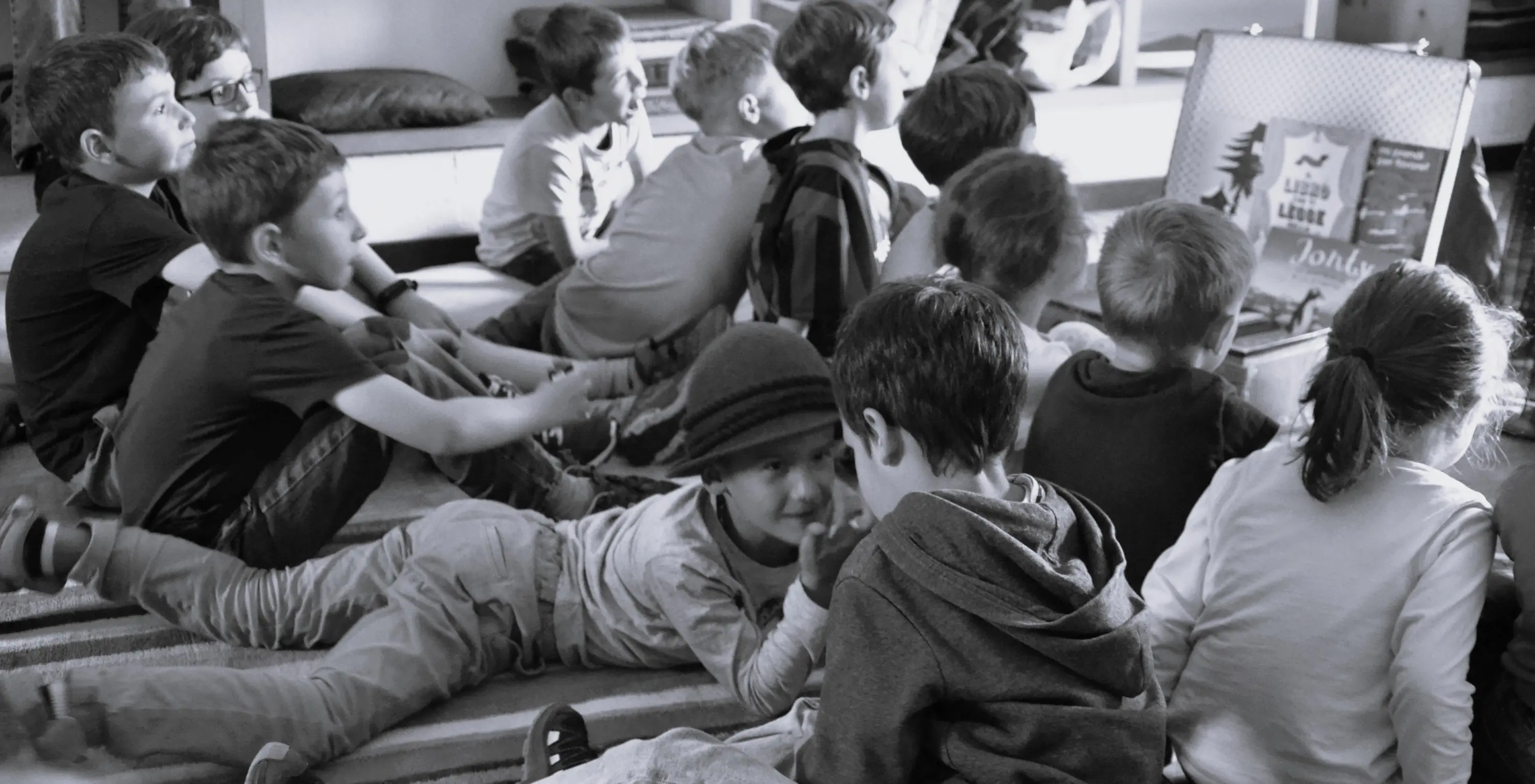 Un gruppo di bambini seduti e distesi su un tappeto, concentrati su un'attività, con alcuni che parlano tra di loro e altri che ascoltano attentamente. L'atmosfera è informale e amichevole.