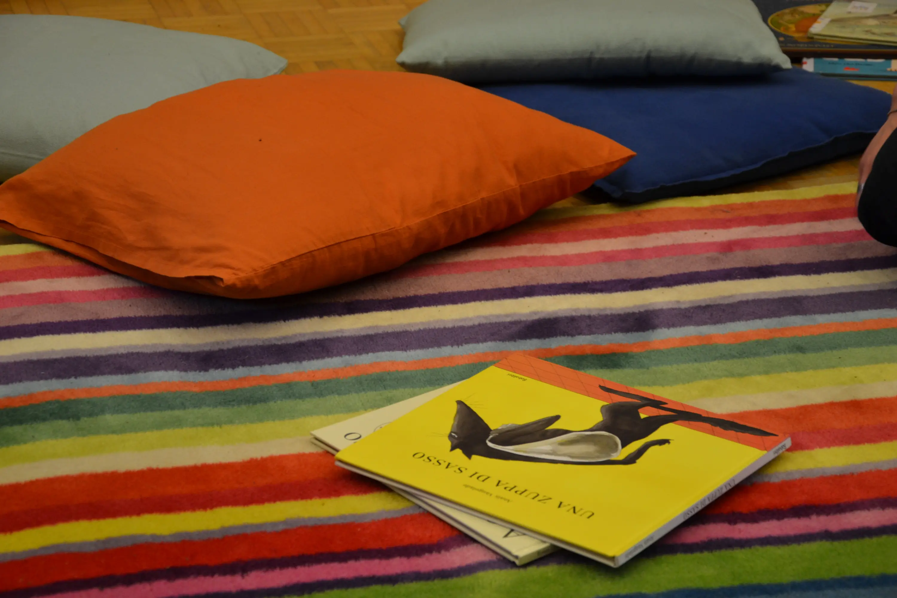 Cuscini colorati disposti su un tappeto a righe, con un libro illustrato aperto sopra di essi.