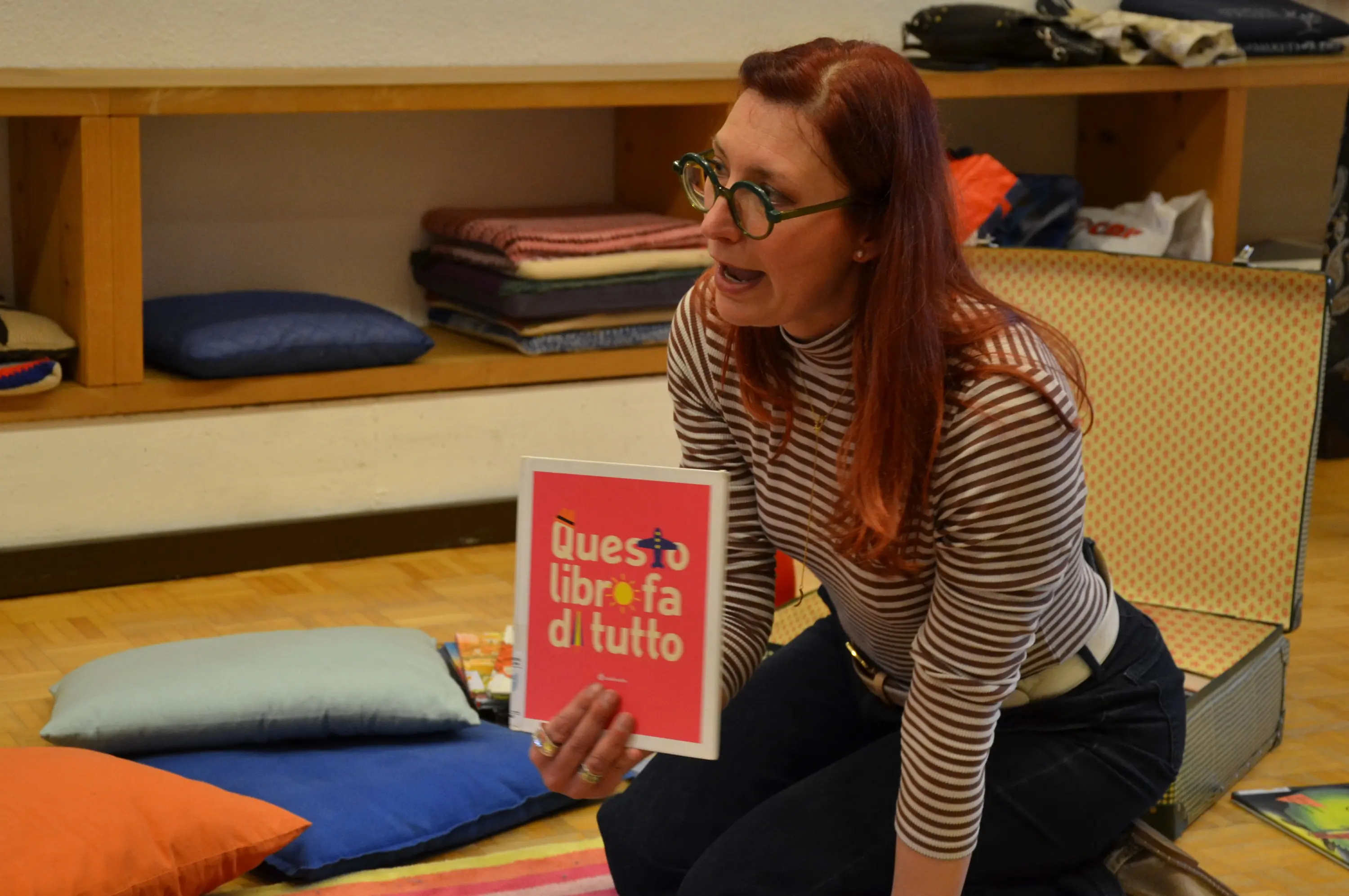 Una donna con occhiali rotondi e capelli rossi tiene un libro colorato in una mano, mentre si china verso un gruppo di cuscini colorati sul pavimento. Indossa una maglietta a righe e sembra impegnata a leggere o spiegare qualcosa. Sullo sfondo si vedono altri oggetti e coperte.