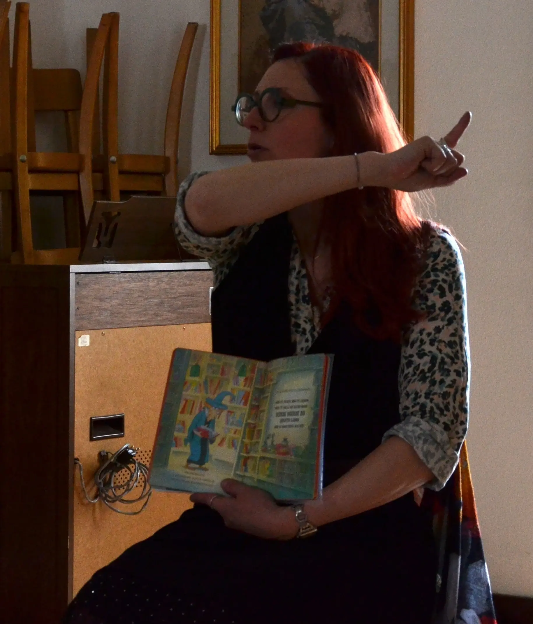 Una donna con capelli rossi e occhiali mostra un libro illustrato, apparentemente spiegando una storia, mentre indica verso un angolo. Sullo sfondo si vedono sedie impilate e una parete.
