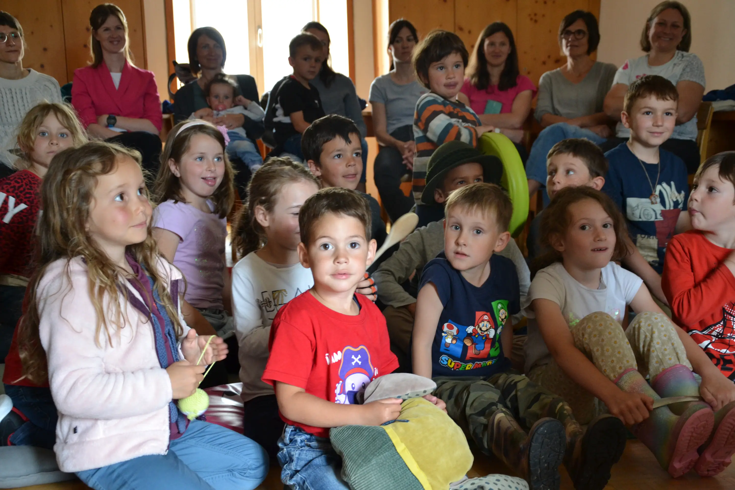 Un gruppo di bambini seduti in un'aula, sorridenti e attenti. Alcuni indossano magliette colorate e altri hanno oggetti in mano. Sullo sfondo, adulti osservano la scena.