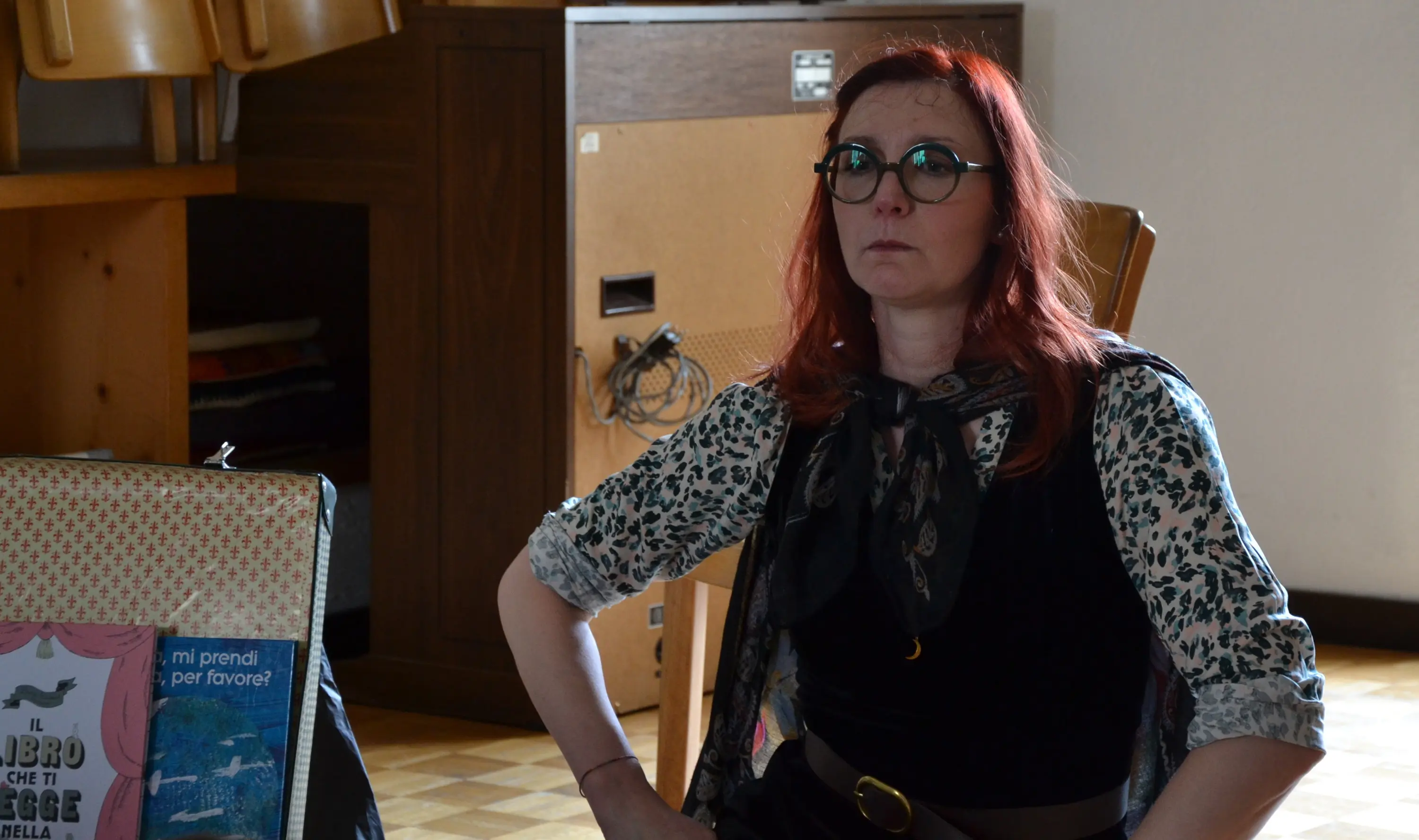 Una donna con capelli rossi e occhiali, seduta in un ambiente interno. Indossa una blusa con fantasia leopardata e una sciarpa nera. Sullo sfondo, si intravedono mobili e una valigia con libri. La sua espressione è seria e pensierosa.