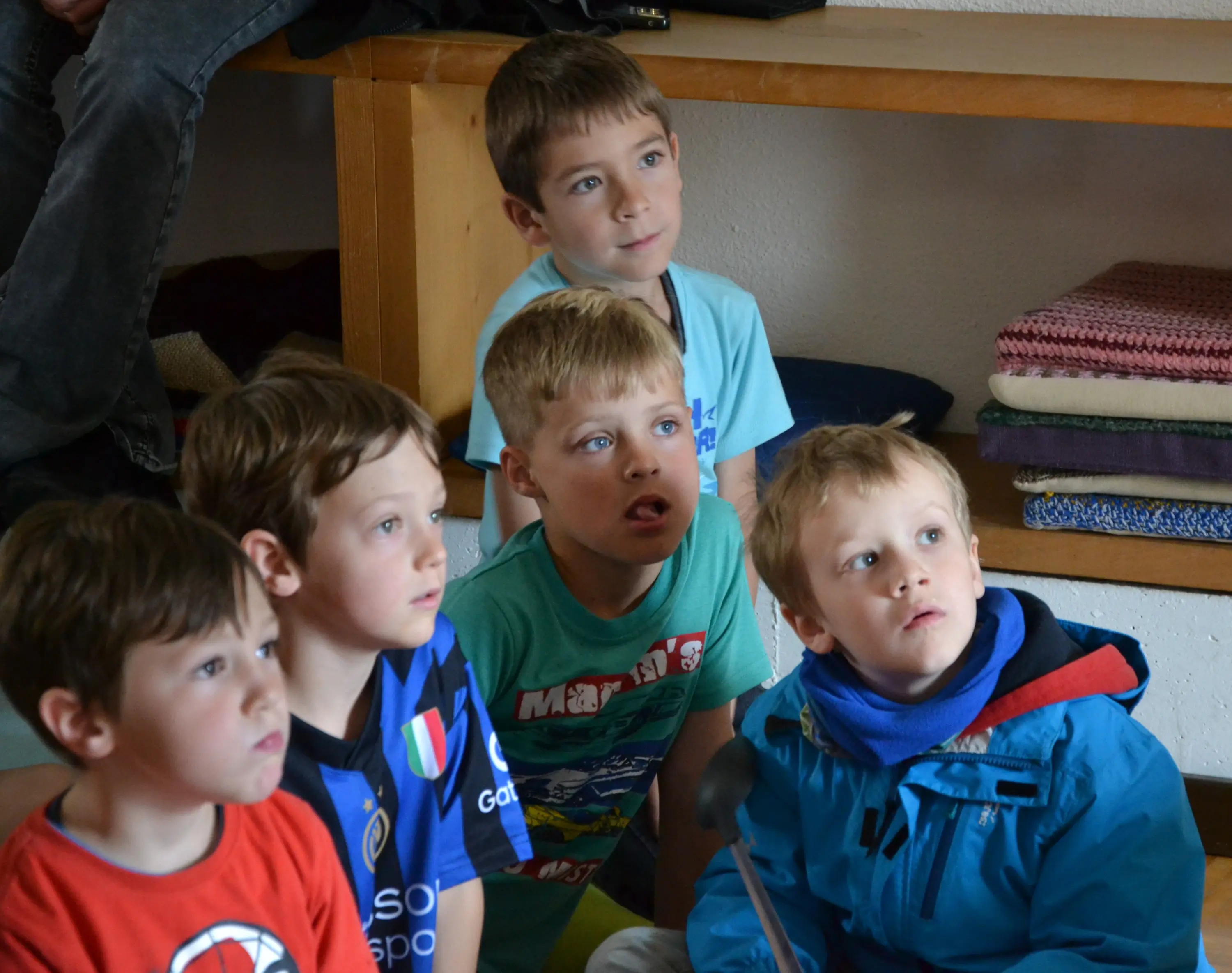 Cinque bambini seduti, intenti a guardare qualcosa con espressioni curiose. Indossano magliette colorate e si trovano in un ambiente luminoso e informale.