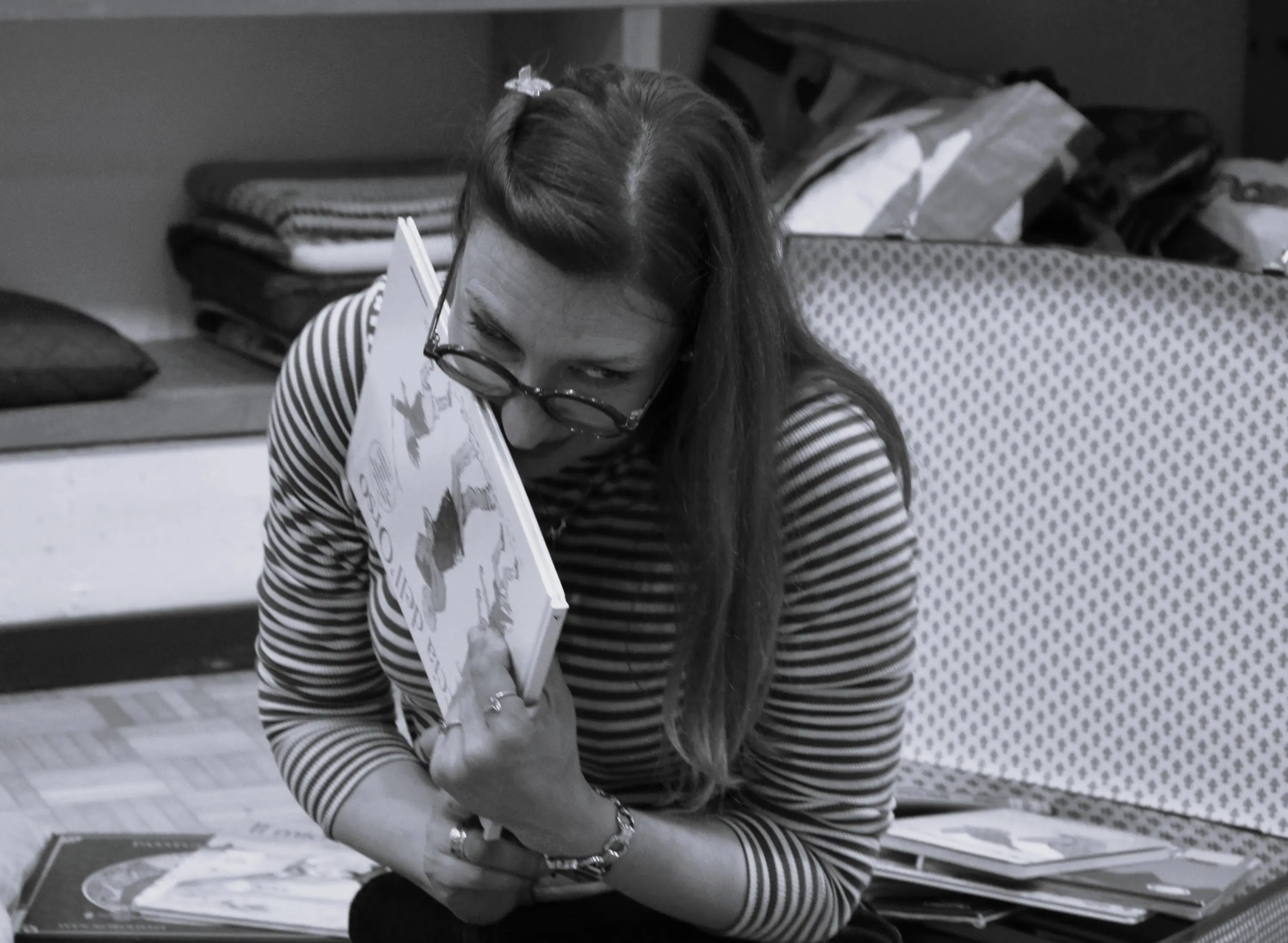 Una donna con occhiali abbraccia un libro, sorridendo mentre si trova su un pavimento rivestito. Indossa una maglietta a righe ed è circondata da libri disposti in modo disordinato.