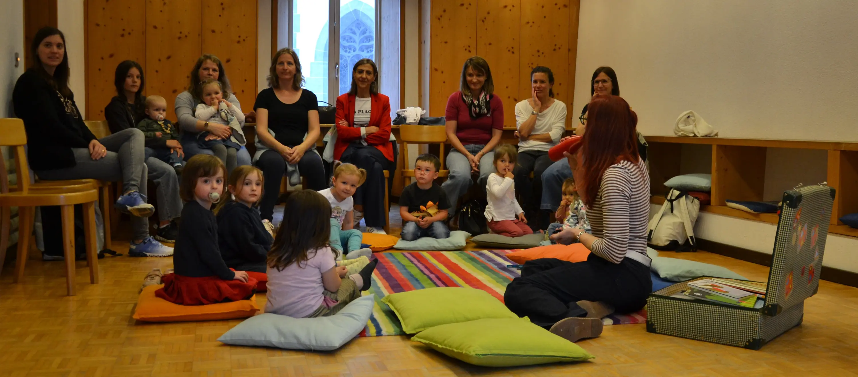 Un gruppo di adulti e bambini seduti su cuscini colorati in una stanza accogliente, mentre una donna con capelli rossi racconta una storia. Le pareti in legno creano un'atmosfera calda e familiare.