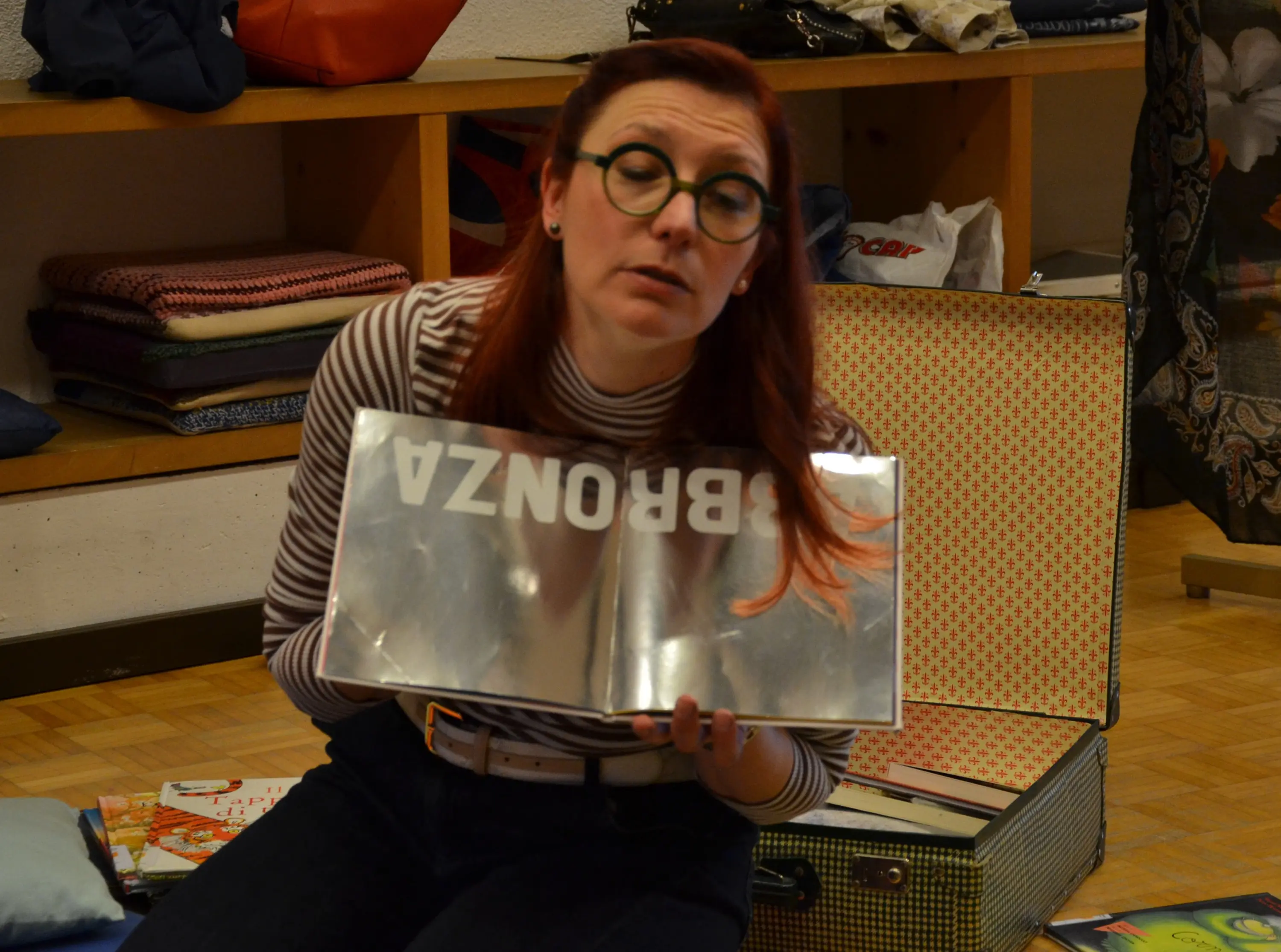 Donna con occhiali rotondi e capelli rossi tiene in mano un libro aperto con la parola "BRONZA" stampata in grande. Sullo sfondo sono visibili scaffali con tessuti e libri.