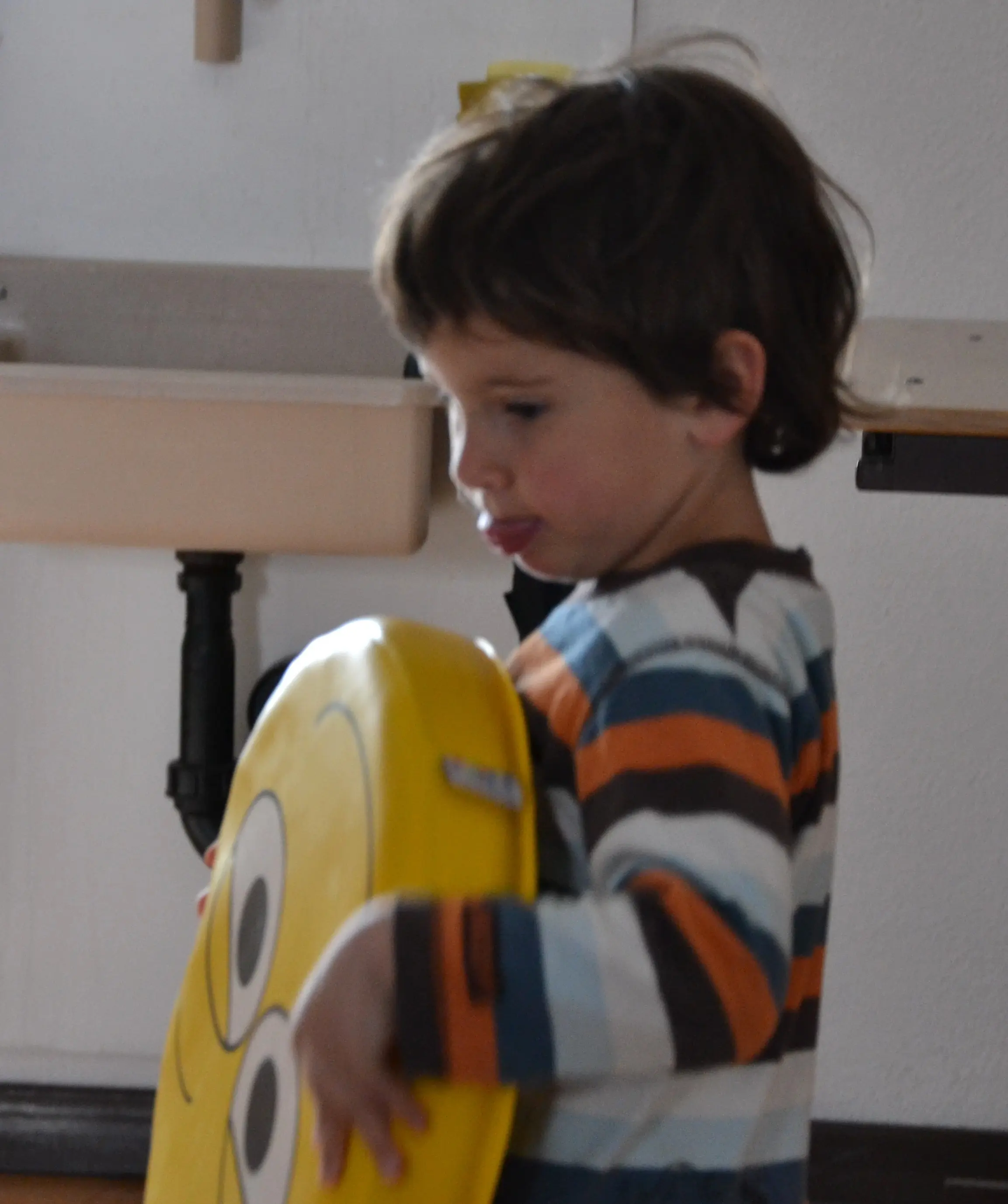 Un bambino con una maglietta a righe colorate tiene in mano un cuscino giallo a forma di sorriso, mostrando una faccia buffa.