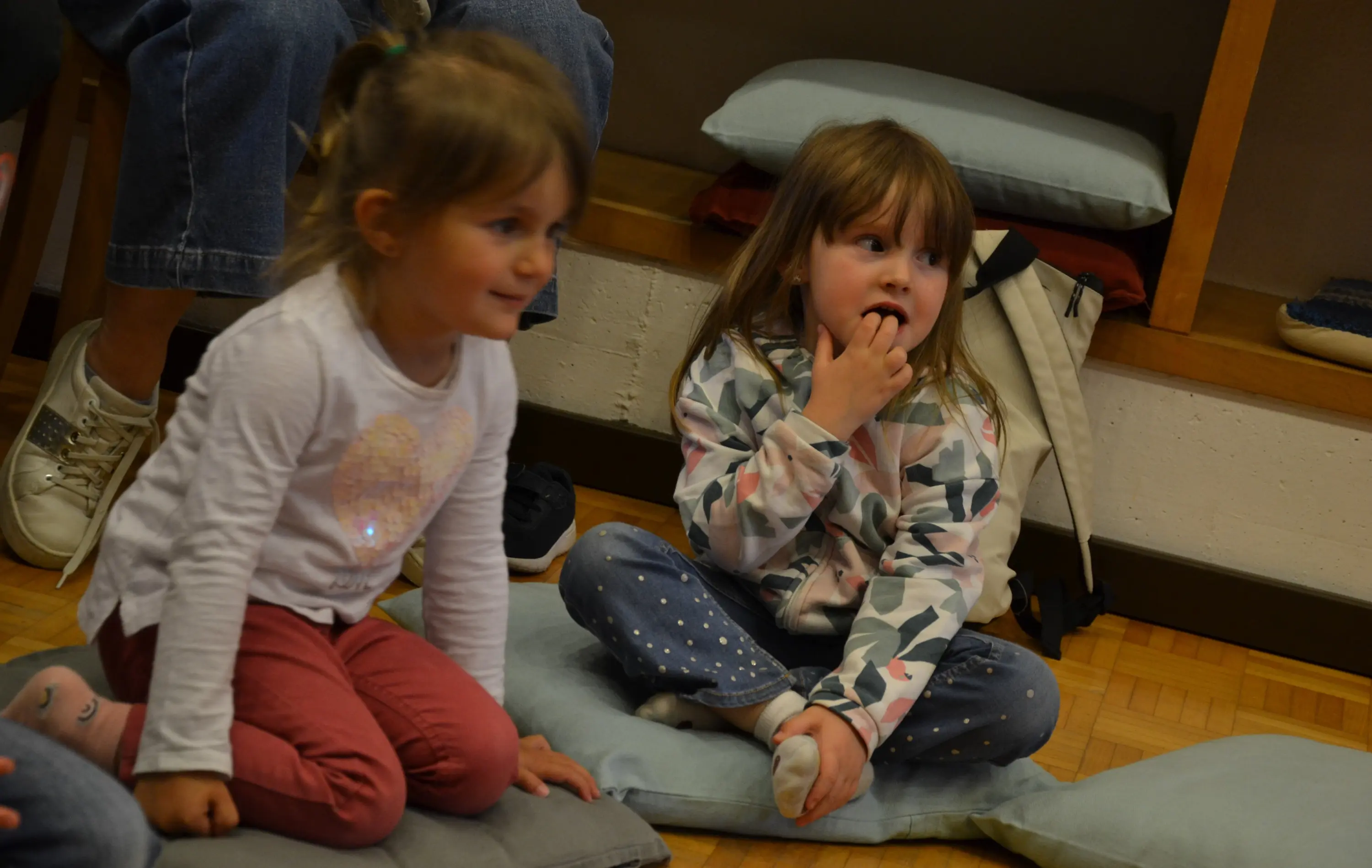 Due bambine sedute su cuscini in un ambiente interno. Una delle bambine indossa una maglietta bianca con un motivo luccicante e pantaloni rosa, mentre l'altra ha una giacca a motivo camo e pantaloni a pois. Entrambe sembrano interessate e sorridenti.