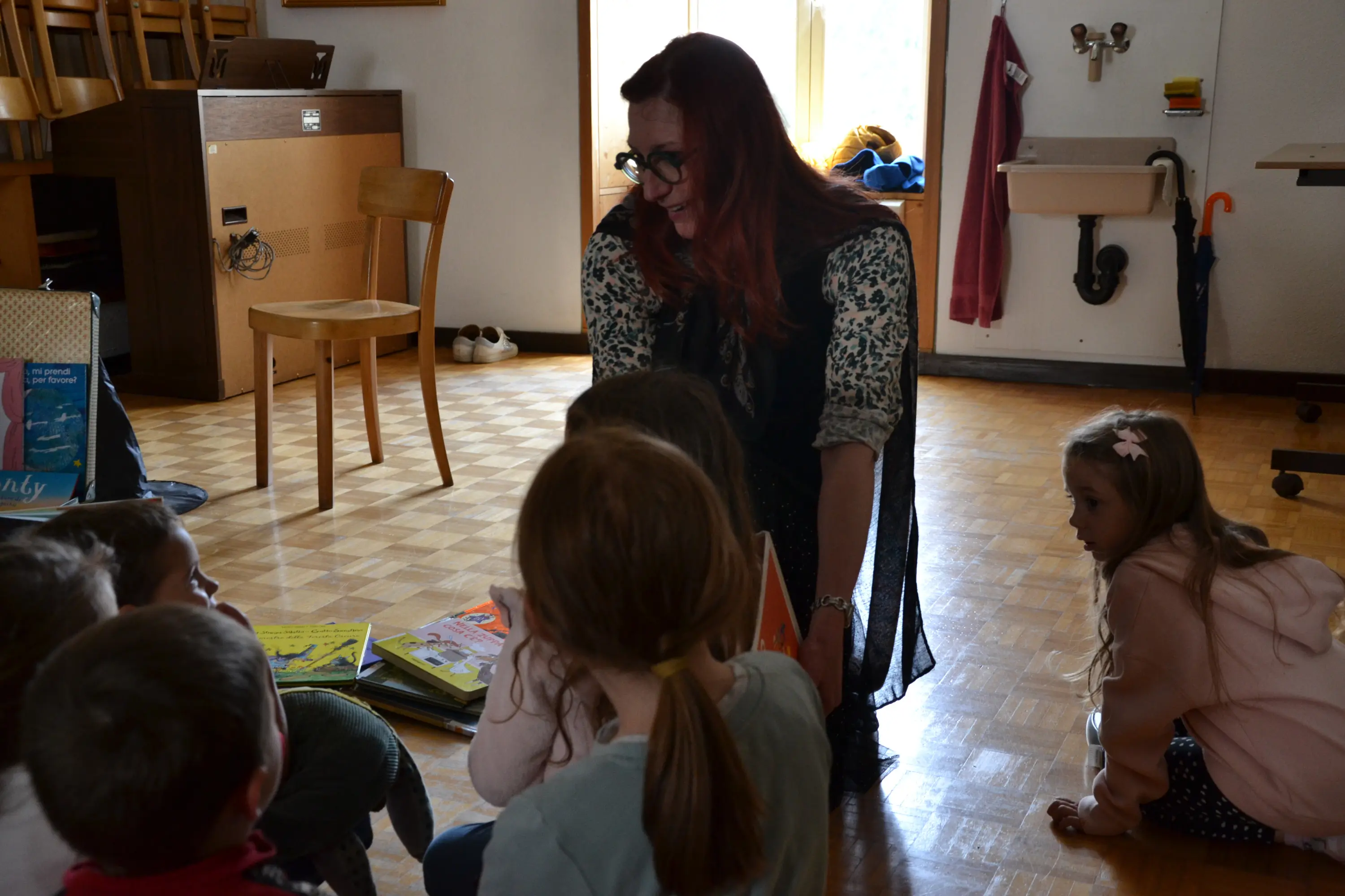 Una donna con capelli rossi e occhiali interagisce con un gruppo di bambini seduti a terra, circondati da libri. L'ambiente è luminoso e accogliente, con un pavimento in legno e alcuni mobili sullo sfondo.
