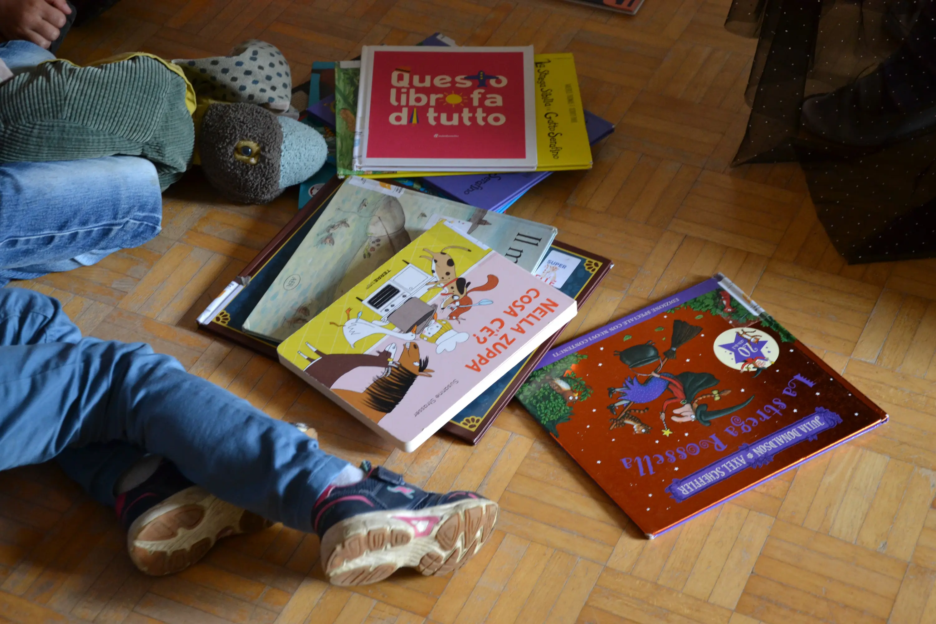 Libri colorati sparsi sul pavimento, con un paio di piedi di un bambino visibili e un peluche in primo piano. I titoli includono "Questo libro fa di tutto" e "Il piccolo principe".