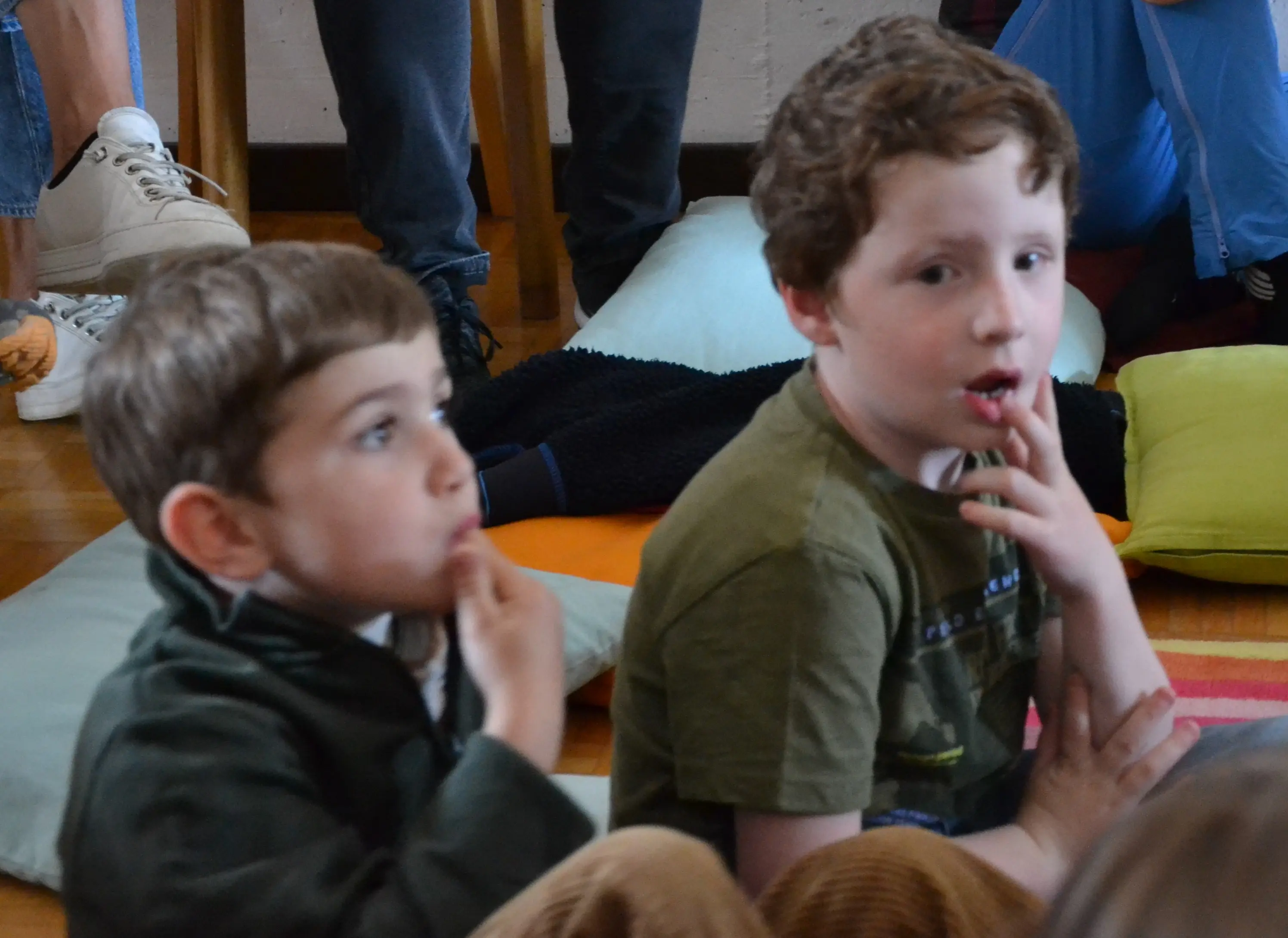 Due bambini seduti su cuscini colorati, apparentemente assorti e pensierosi. Uno ha i capelli castani e corti, l'altro ha i capelli ricci e rossi. Entrambi si toccano le labbra con le dita.