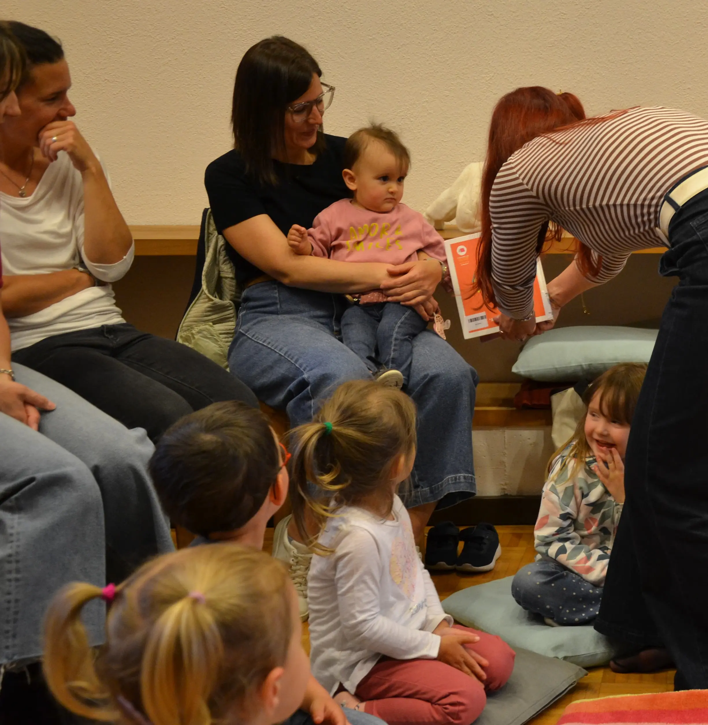 Un gruppo di adulti e bambini seduti in una stanza. Una donna in rosso mostra un libro a un piccolo gruppo di bambini, mentre una madre tiene in braccio una bambina. L’atmosfera è conviviale e coinvolgente.