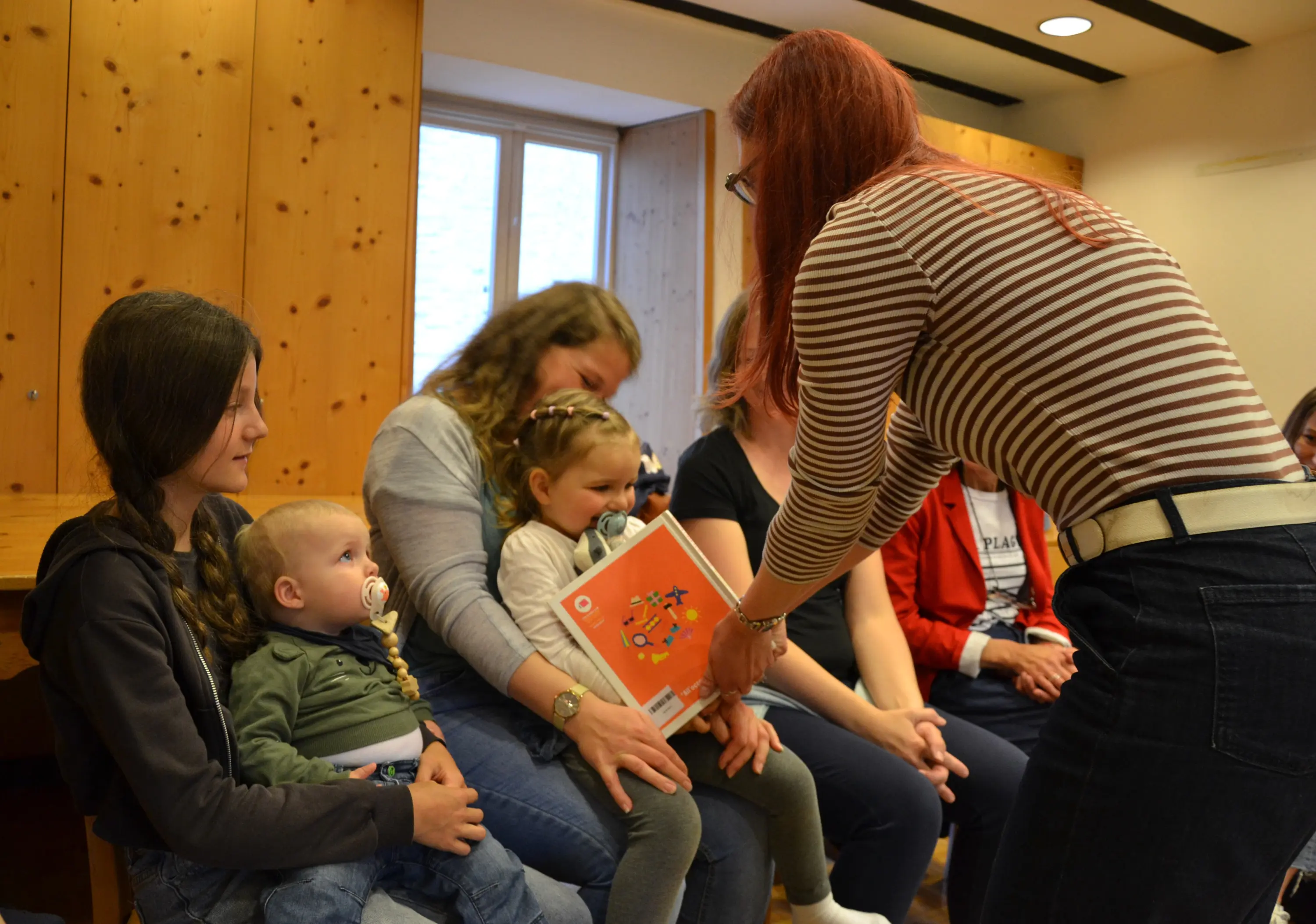 Un gruppo di adulti e bambini seduti in una stanza. Una donna con capelli rossi sta consegnando un libro a una bambina seduta accanto a un'altra donna. I bambini sembrano interessati e alcuni hanno ciucci.