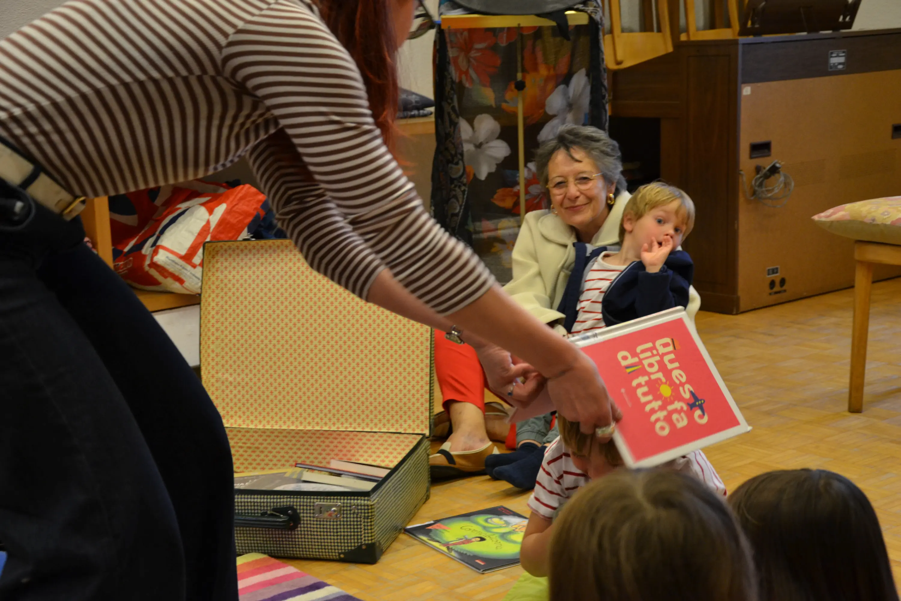 Un adulto porge un libro a un bambino mentre un'altra donna e altri bambini guardano. La scena è luminosa e informale, con oggetti e tappeti colorati sul pavimento.