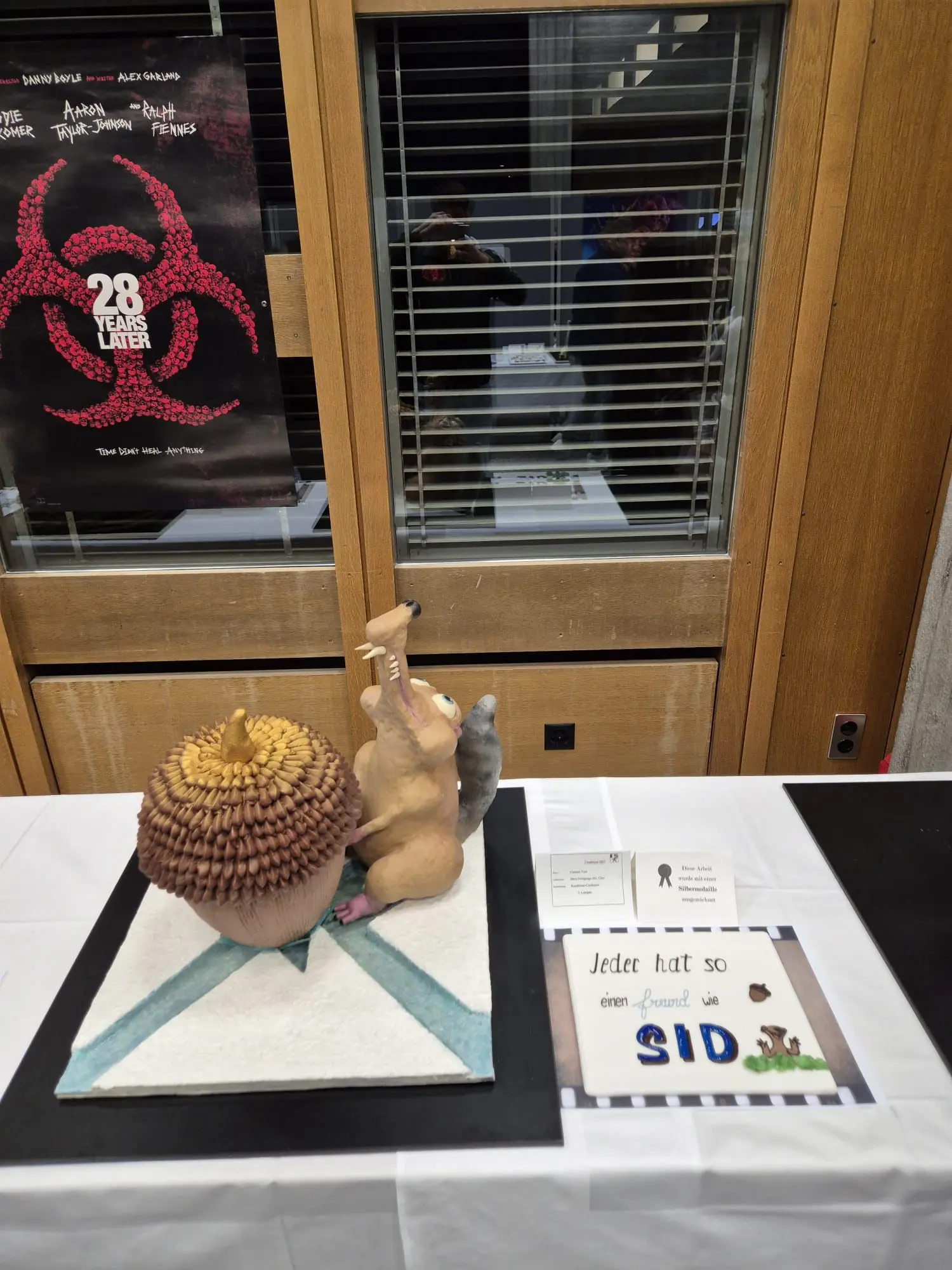 Scultura di un scoiattolo che tiene un grande riccio, esposta su un tavolo con un cartello che indica "Ogniuno ha un amico". Sullo sfondo, un poster di "28 anni dopo" è visibile.