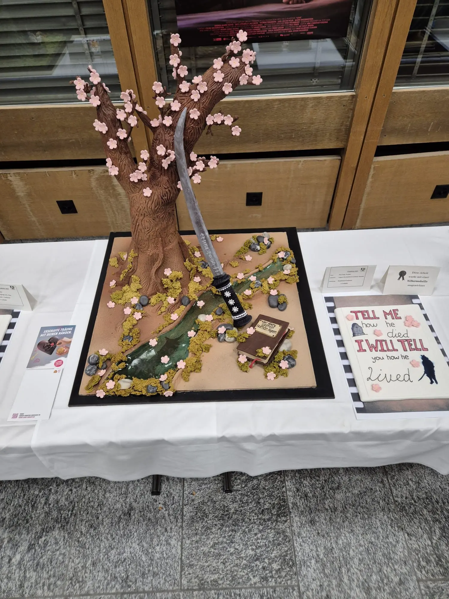 Torta decorata che rappresenta un albero di ciliegio con fiori rosa, una katana, ciottoli e una stele. Sul tavolo ci sono anche cartelli con scritte.