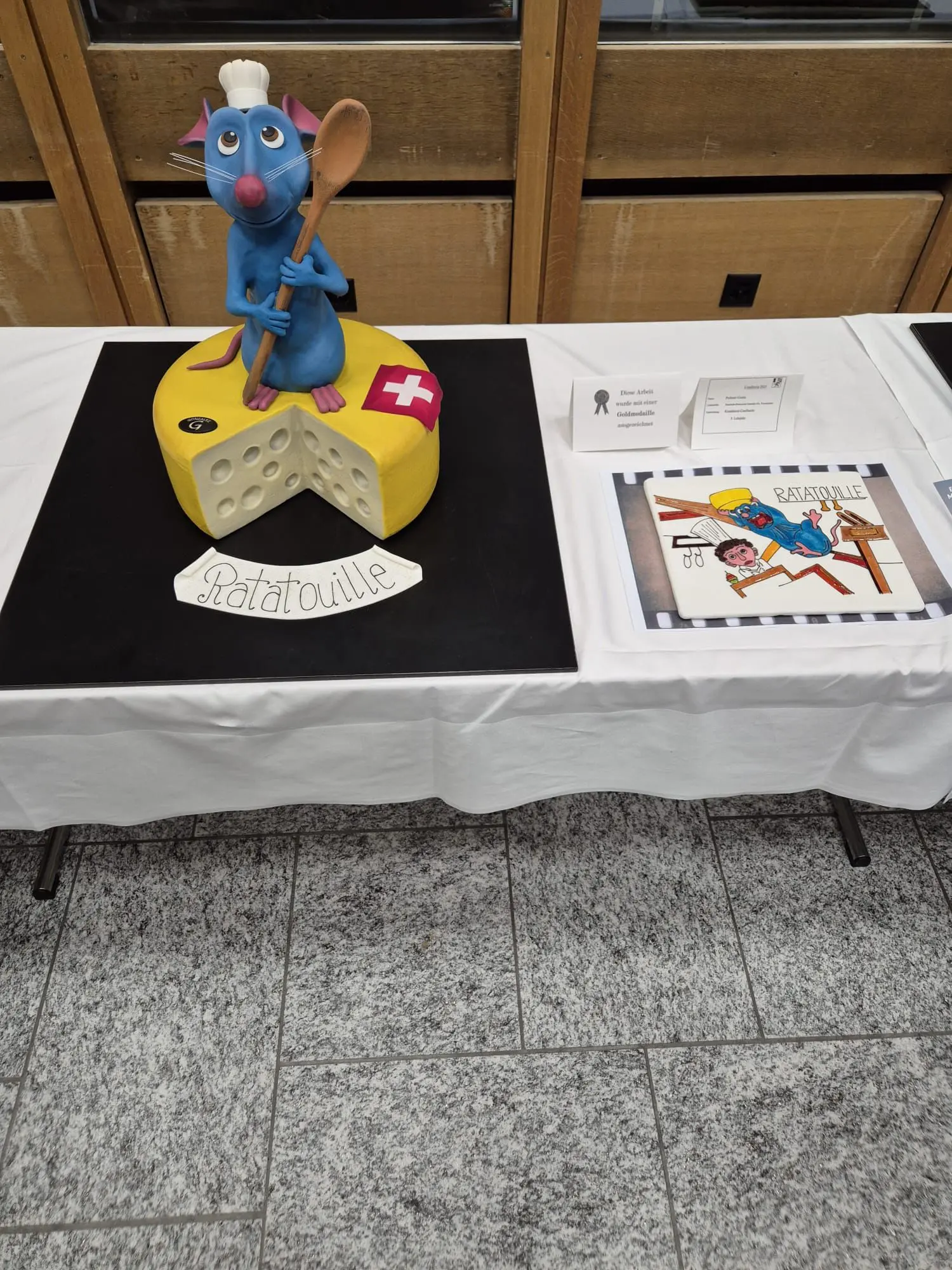 Un grande cake decorato che rappresenta Remy, il topo chef di "Ratatouille", con un cucchiaio in mano e un formaggio giallo con una croce rossa. Accanto, un'opera di arte legata al film.