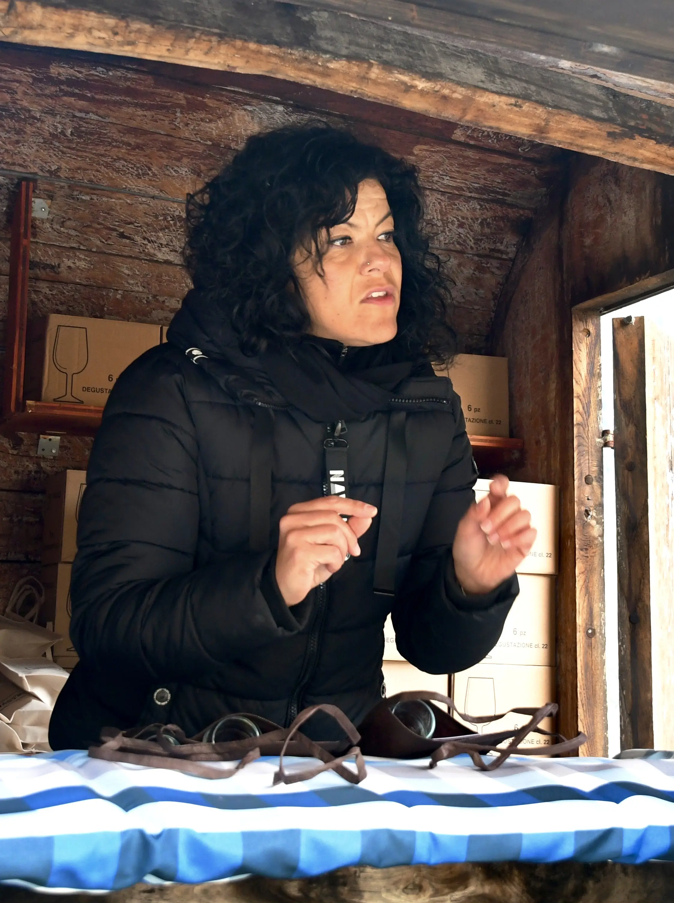 Una donna con capelli ricci e neri indossa un giubbotto nero mentre parla appassionatamente, gesticolando con le mani. Sul tavolo davanti a lei, ci sono degli